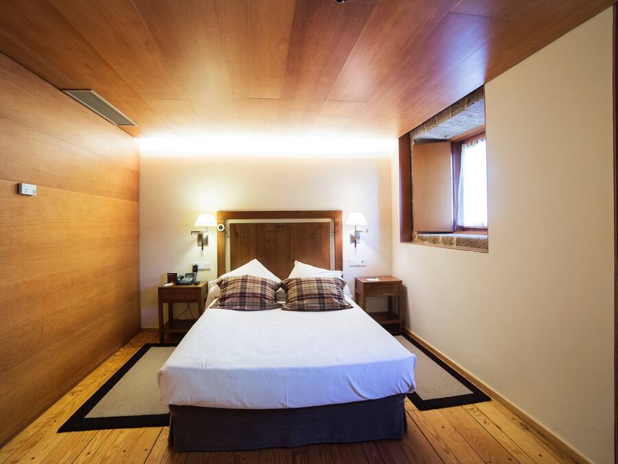 Un dormitorio con techo de madera, una cama con ropa de cama blanca, dos mesitas de noche con lámparas y una ventana con vistas.