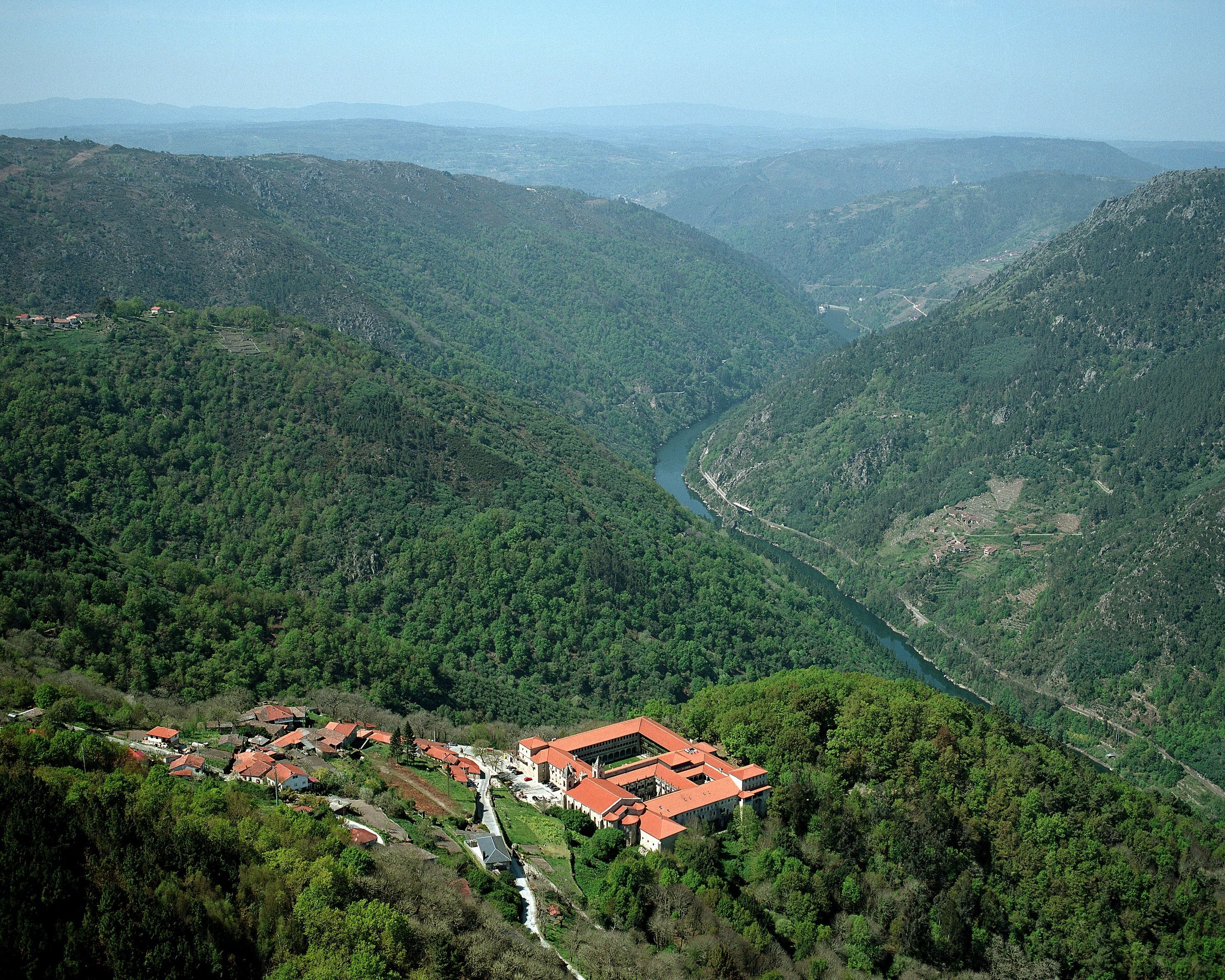 Un paisaje montañoso con un río, un pequeño pueblo y un edificio grande con tejados rojos.
