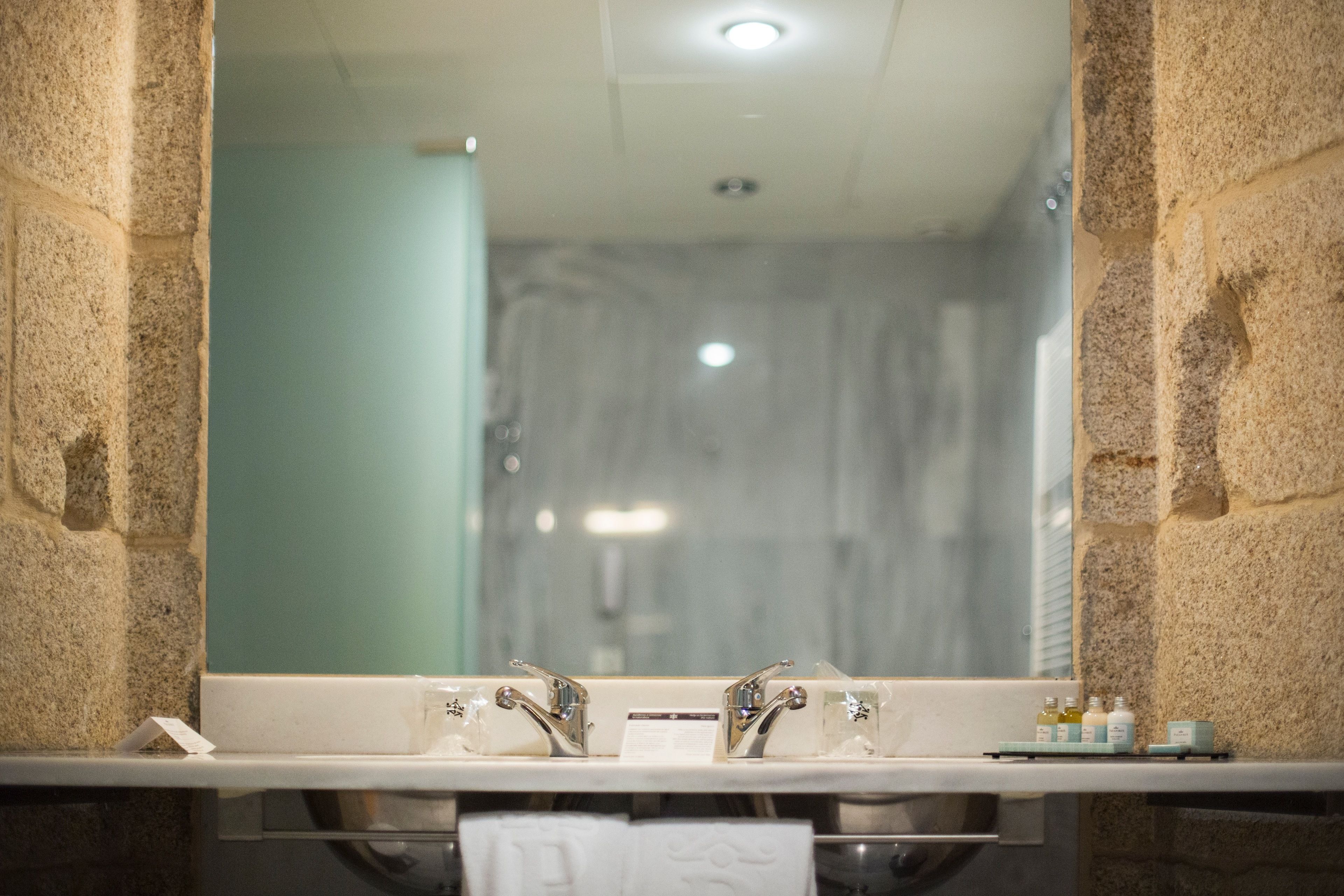 A bathroom with a stone wall, a mirror, a sink with two faucets, and a towel hanging on the wall.