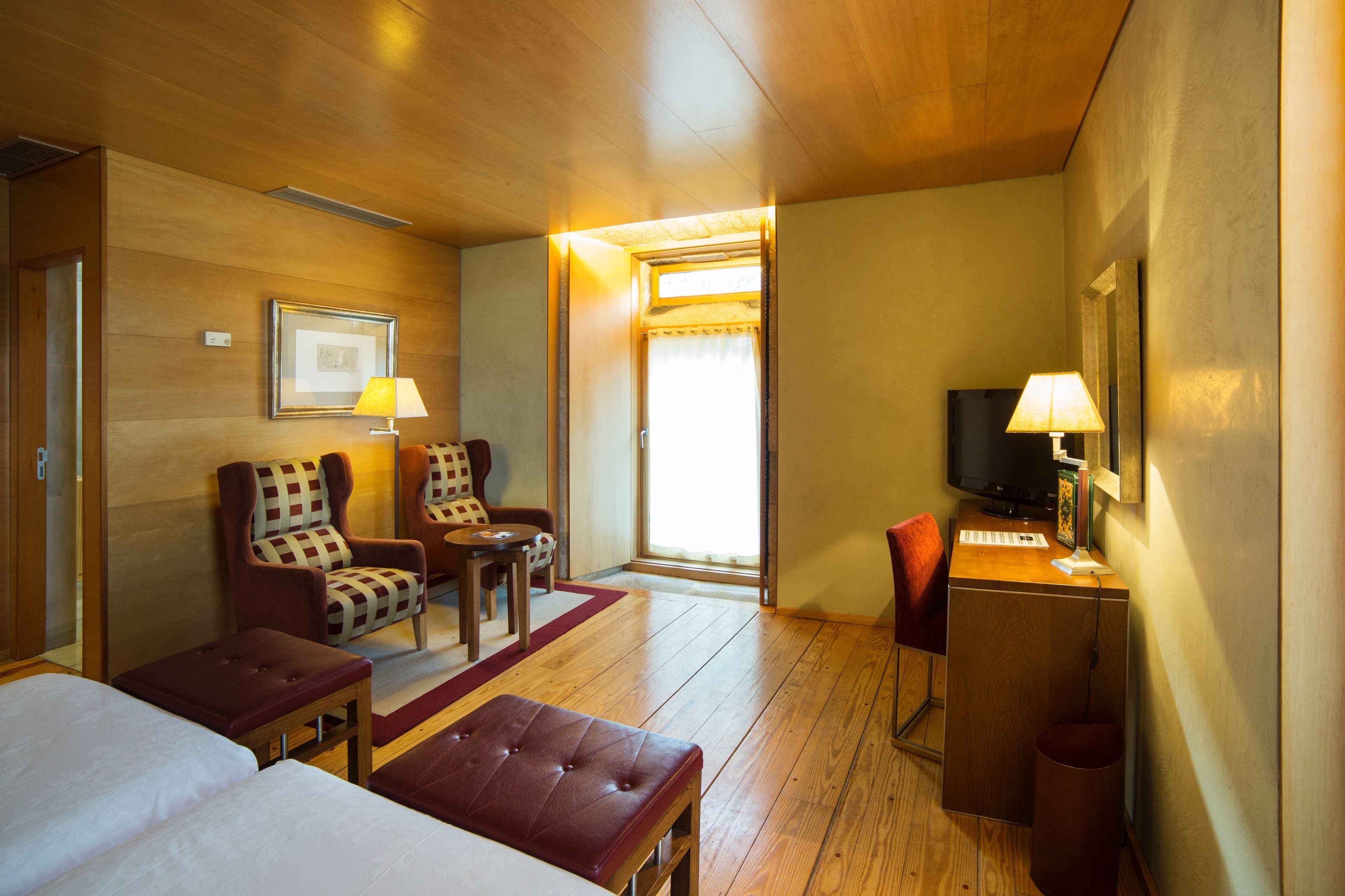 A hotel room with wooden flooring, a bed, two armchairs, a desk with a lamp, a television, and a window with curtains.