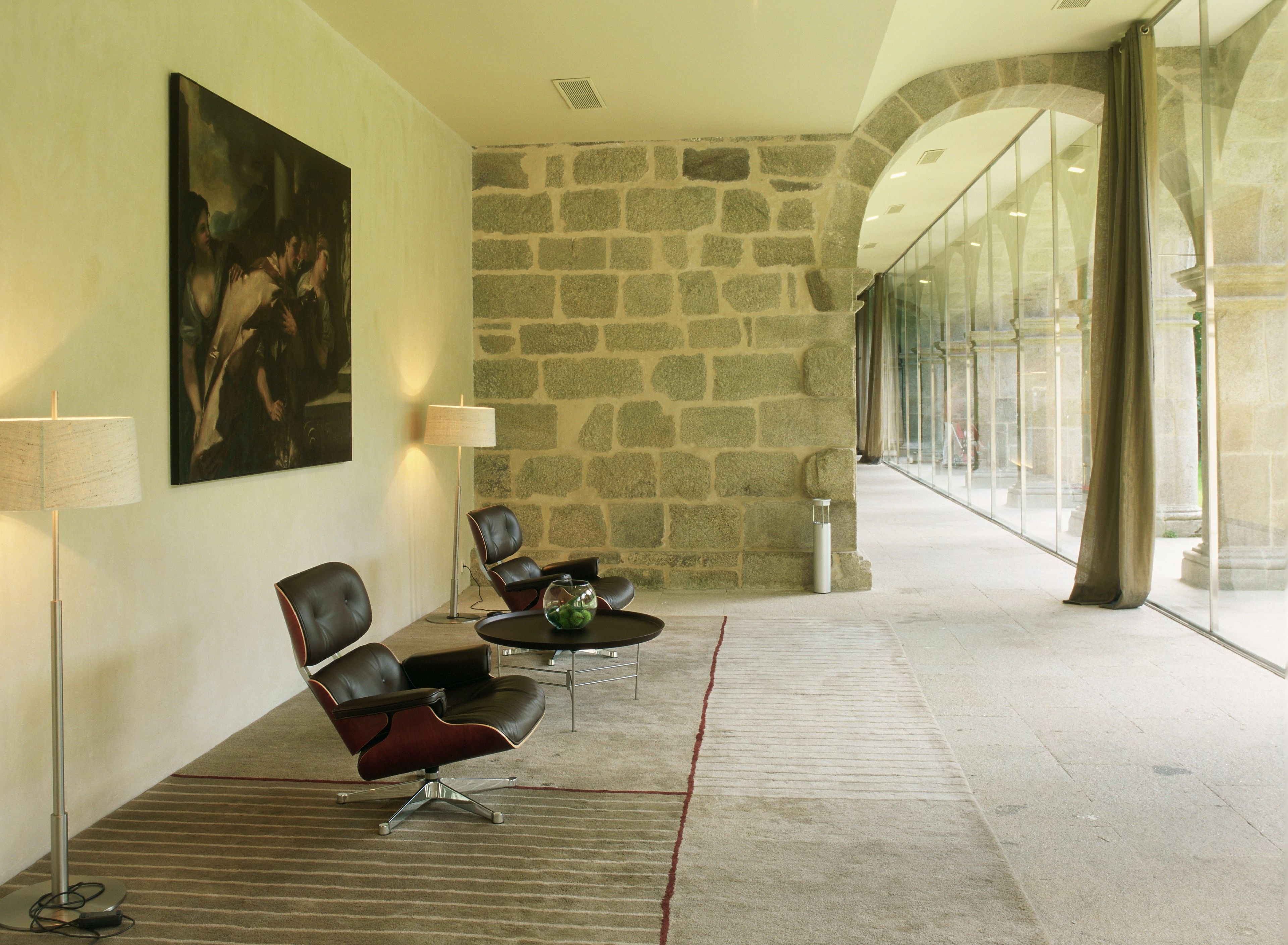 A lobby with stone walls, a large painting, leather chairs, a glass table, and a lamp.