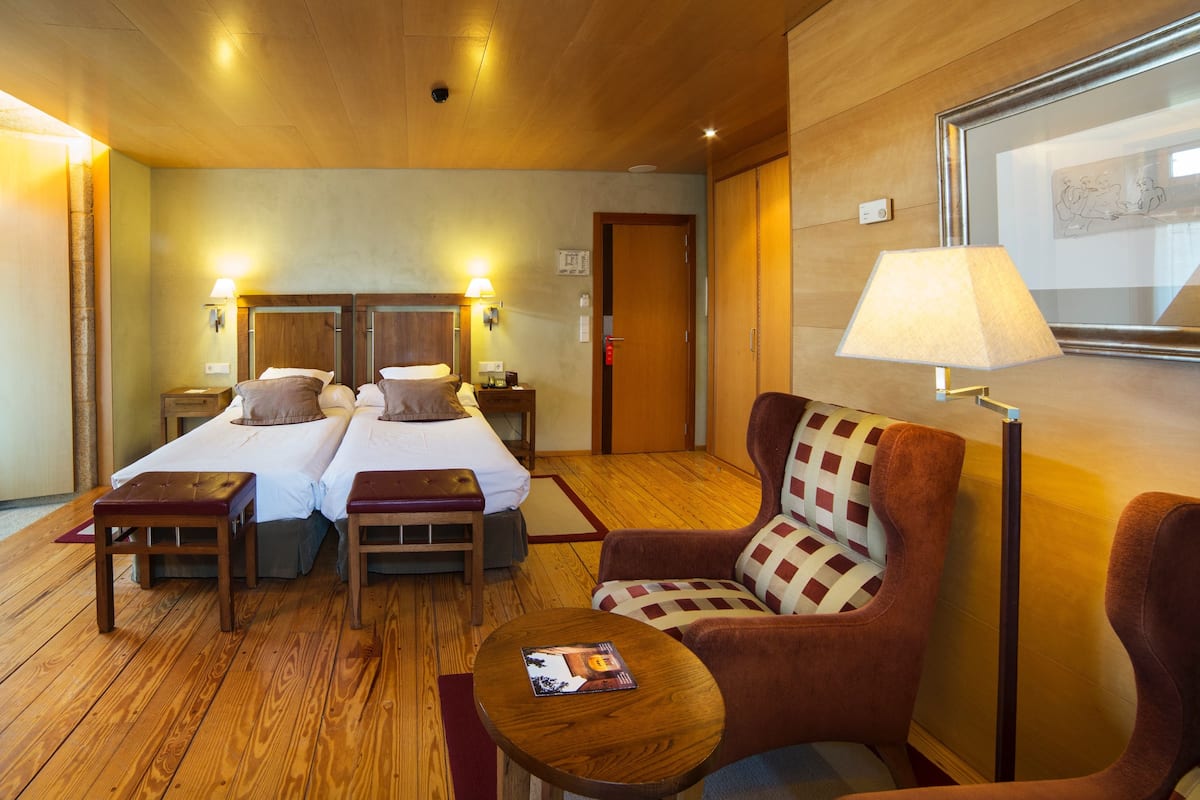 Ein Hotelzimmer mit zwei Betten, Holzboden, einem karierten Sessel, einem kleinen Tisch und einer Lampe.