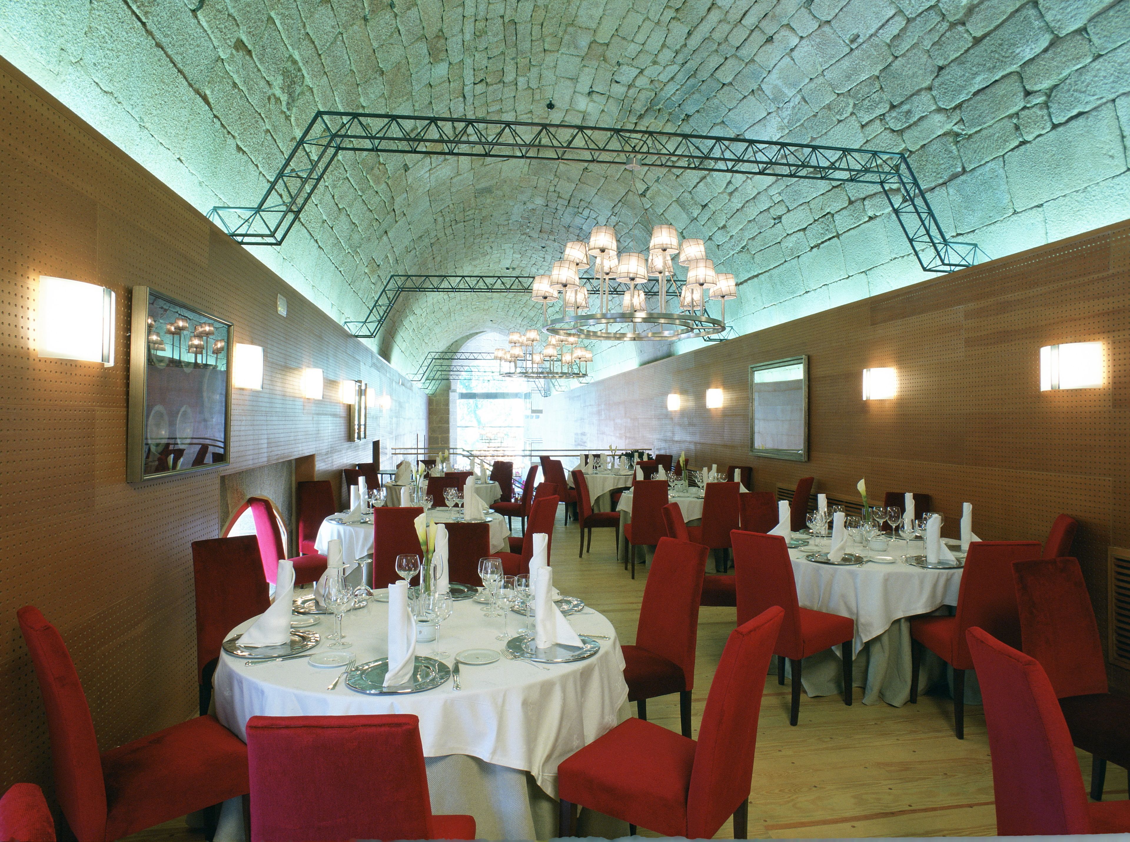 A spacious dining hall with high ceilings and a modern chandelier. Tables are set with white tablecloths and red chairs.