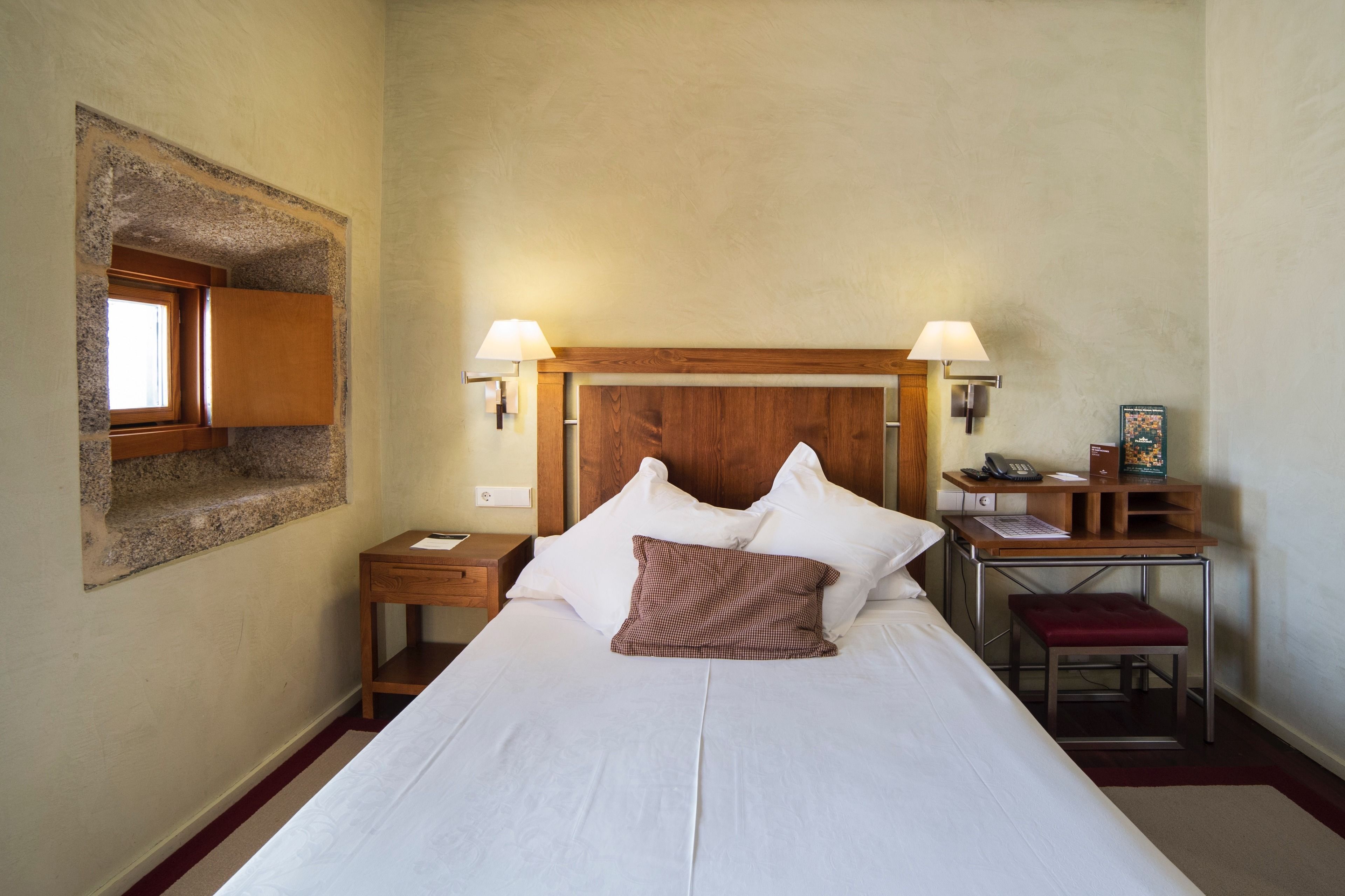 Ein Hotelzimmer mit einem Bett, zwei Nachttischlampen, einem Holznachttisch, einem Schreibtisch mit Stuhl und einem kleinen Fenster.