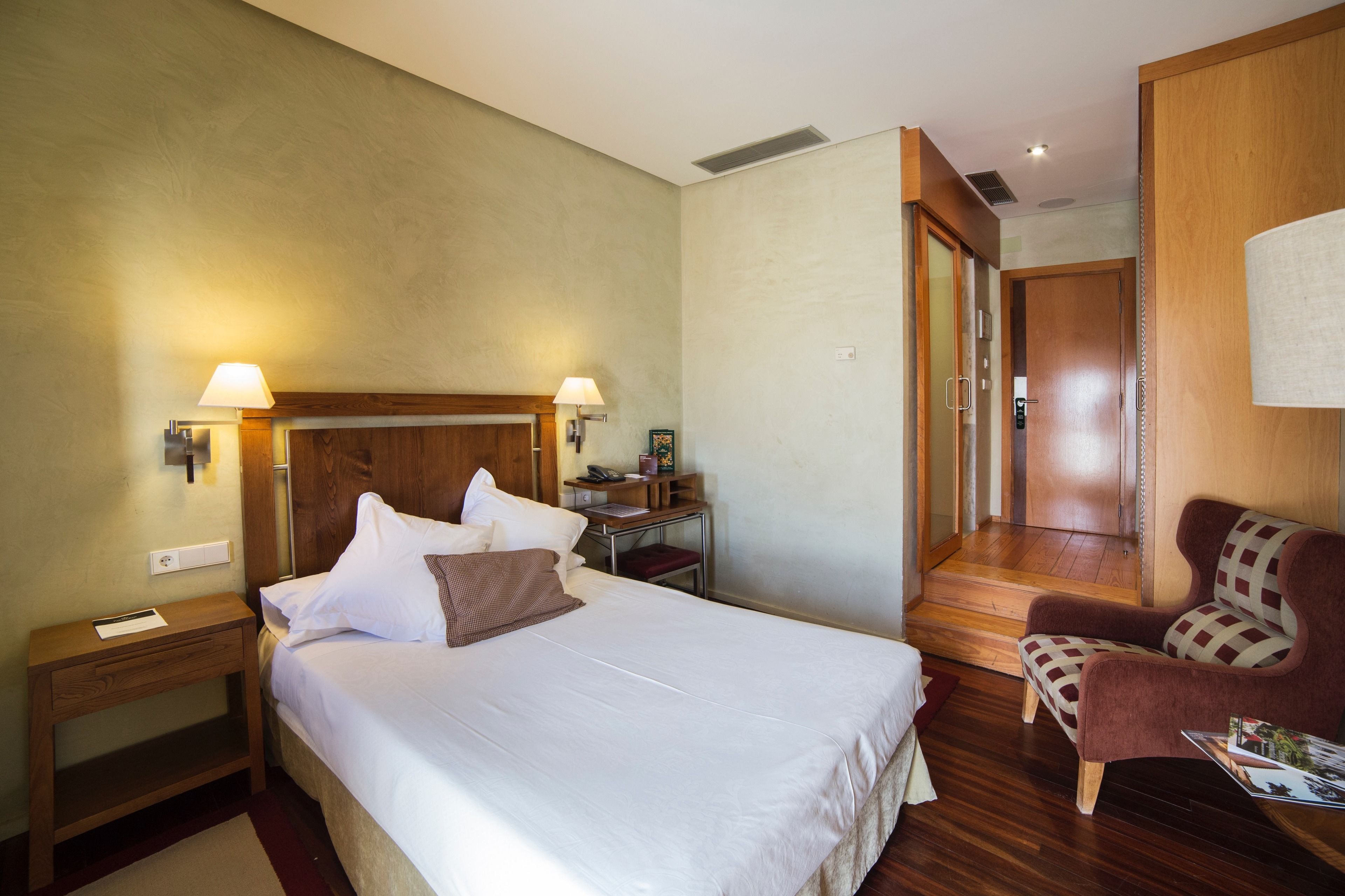 A hotel room with a large bed, bedside tables, a desk, a chair, and a door leading to another room.