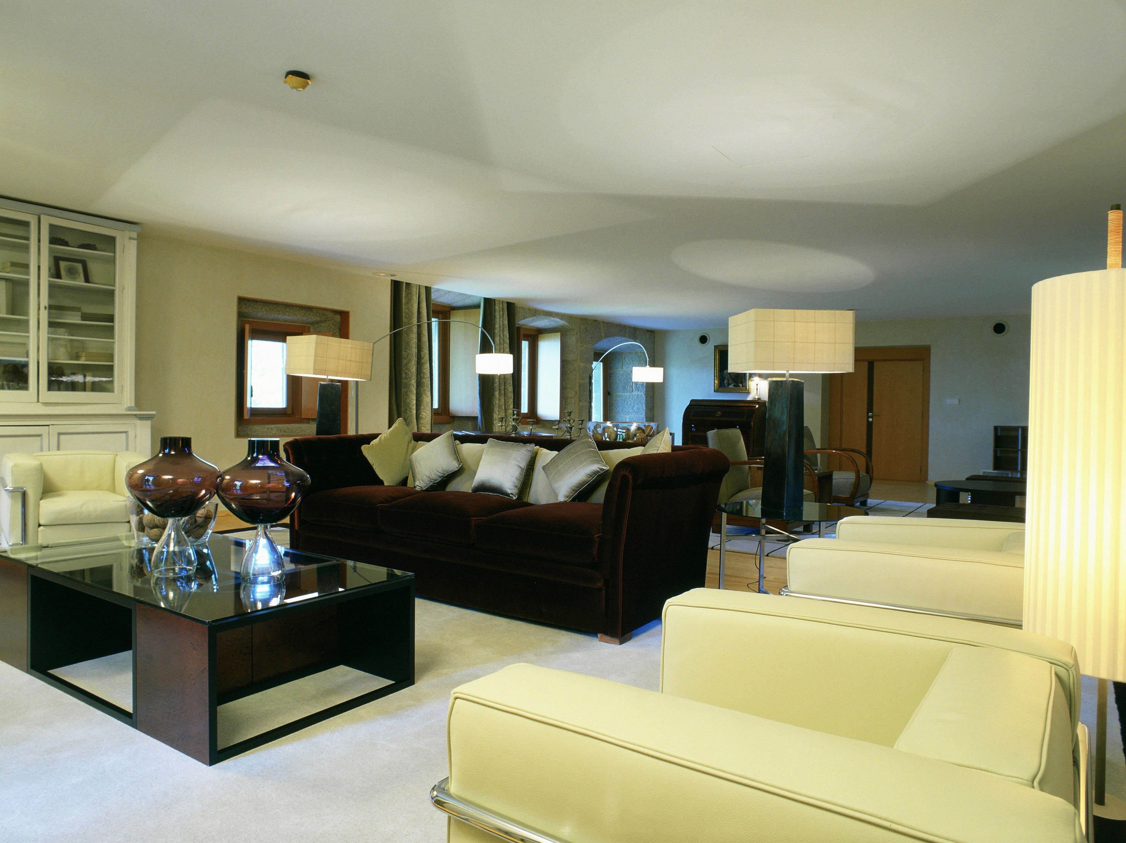 Un salón con un sofá marrón oscuro, dos sillones color crema, una mesa baja de vidrio y una mesa auxiliar de madera oscura. Hay dos lámparas a cada lado del sofá y un espejo grande encima.
