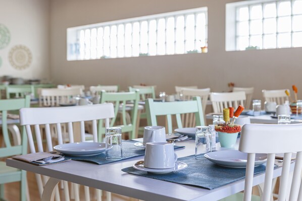Comedor con mesas dispuestas para el servicio, con sillas blancas y manteles individuales azules.
