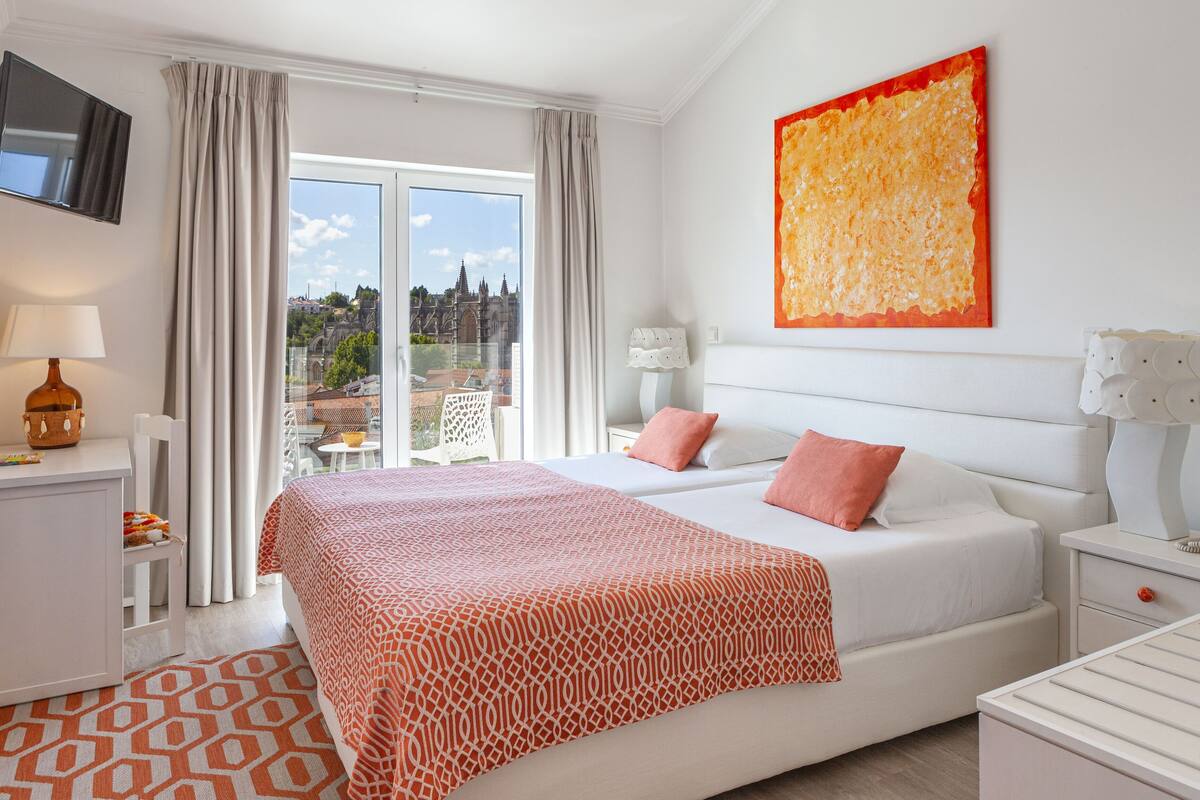 A hotel room with a large bed, two pillows, a nightstand, a lamp, a TV, and a window with a view of a cityscape.