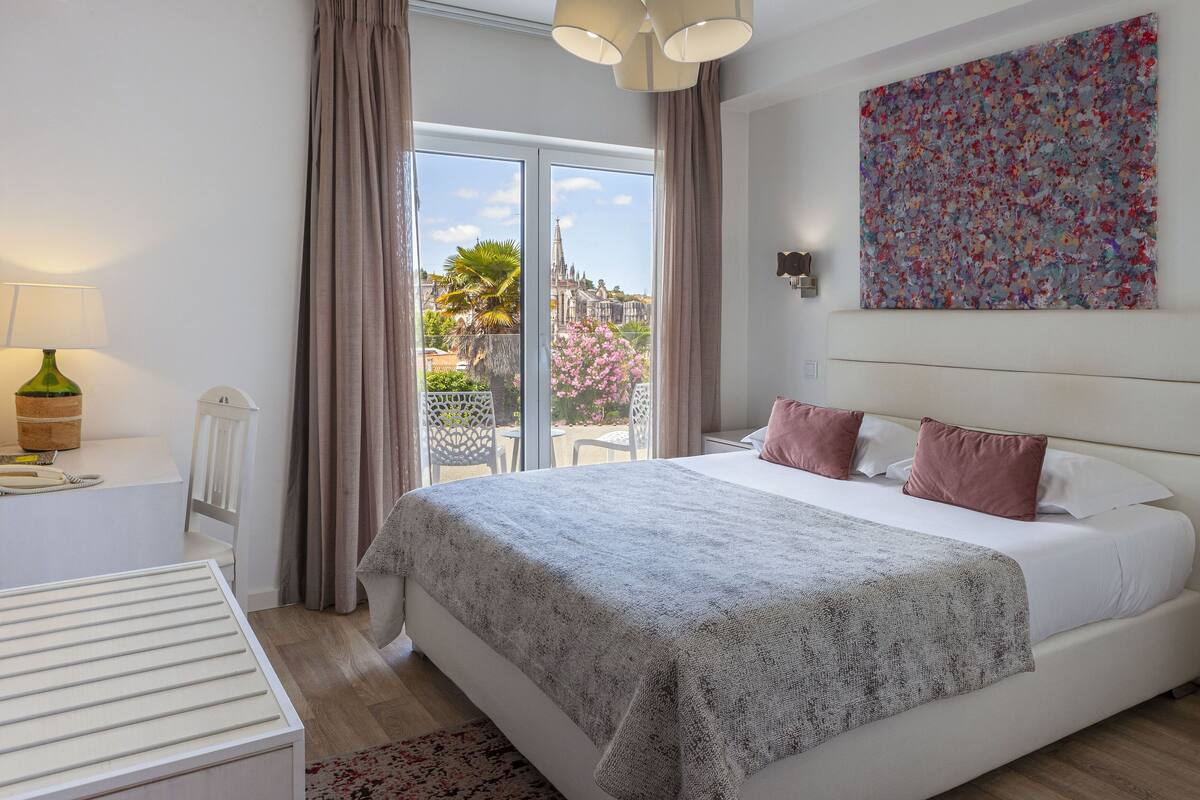 A bedroom with a large bed, a window with curtains, a painting on the wall, and a lamp on a bedside table.