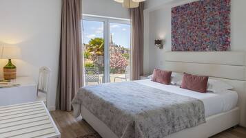 A hotel room with a large bed, a desk with a lamp, a painting on the wall, and a view of a garden and building outside.