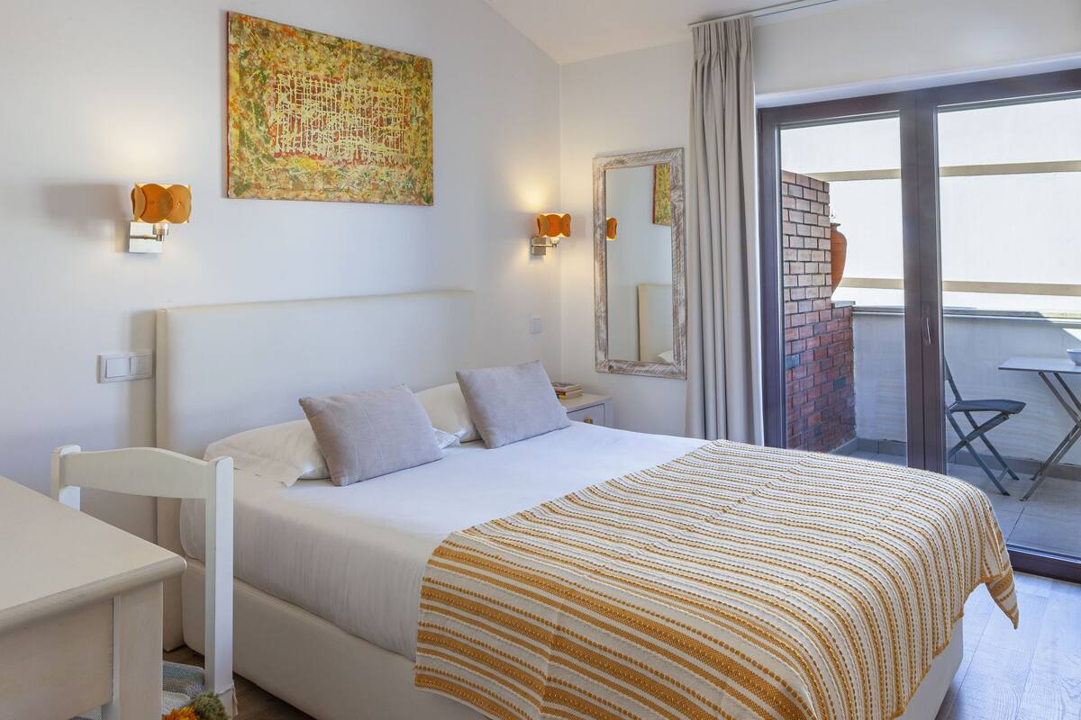 A hotel room with a bed, a desk, a chair, a balcony with a table, and a painting on the wall.