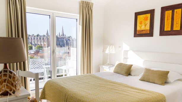 Habitación de hotel con una cama grande, una mesita de noche, una lámpara y vistas a un paisaje urbano a través de la ventana.
