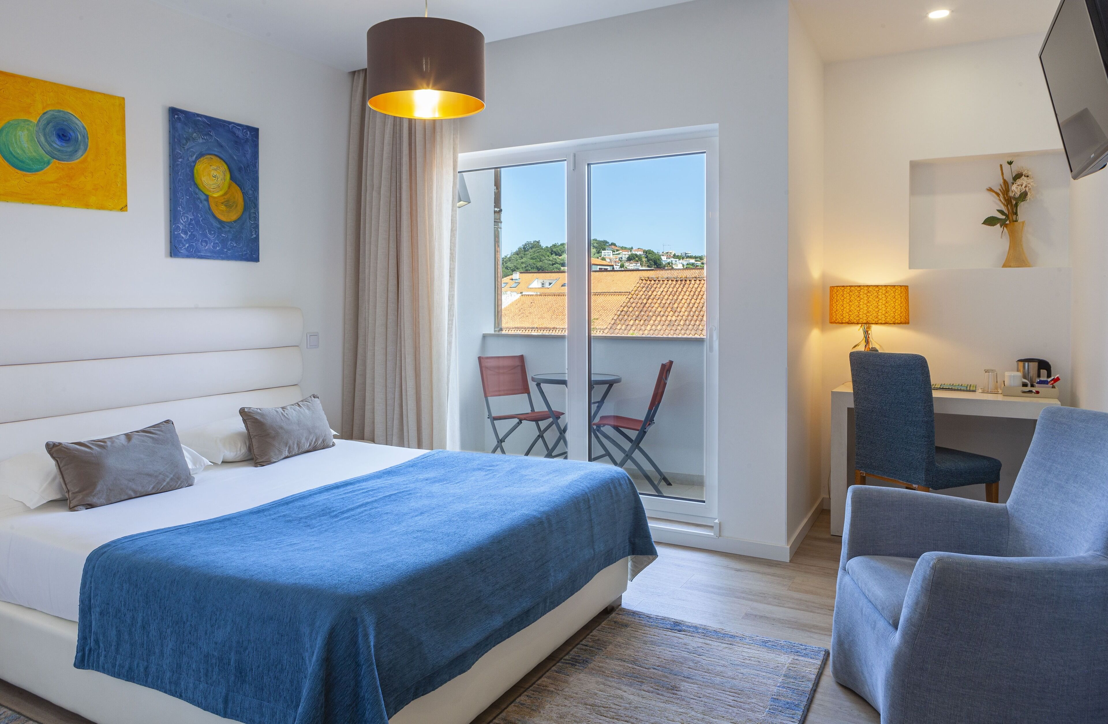 A hotel room with a bed, a desk, a chair, a lamp, and a view of the outside.