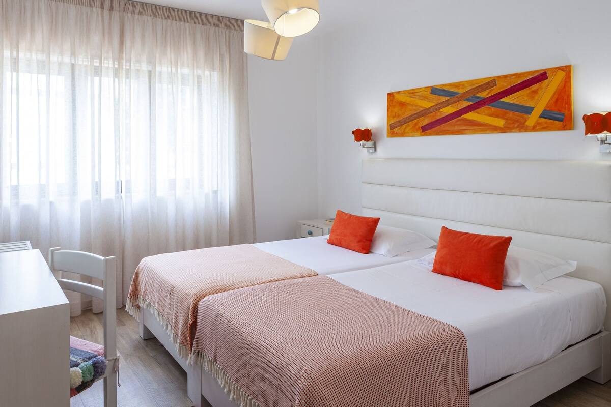 A hotel room with two beds, a desk, a chair, and a large window with sheer curtains.