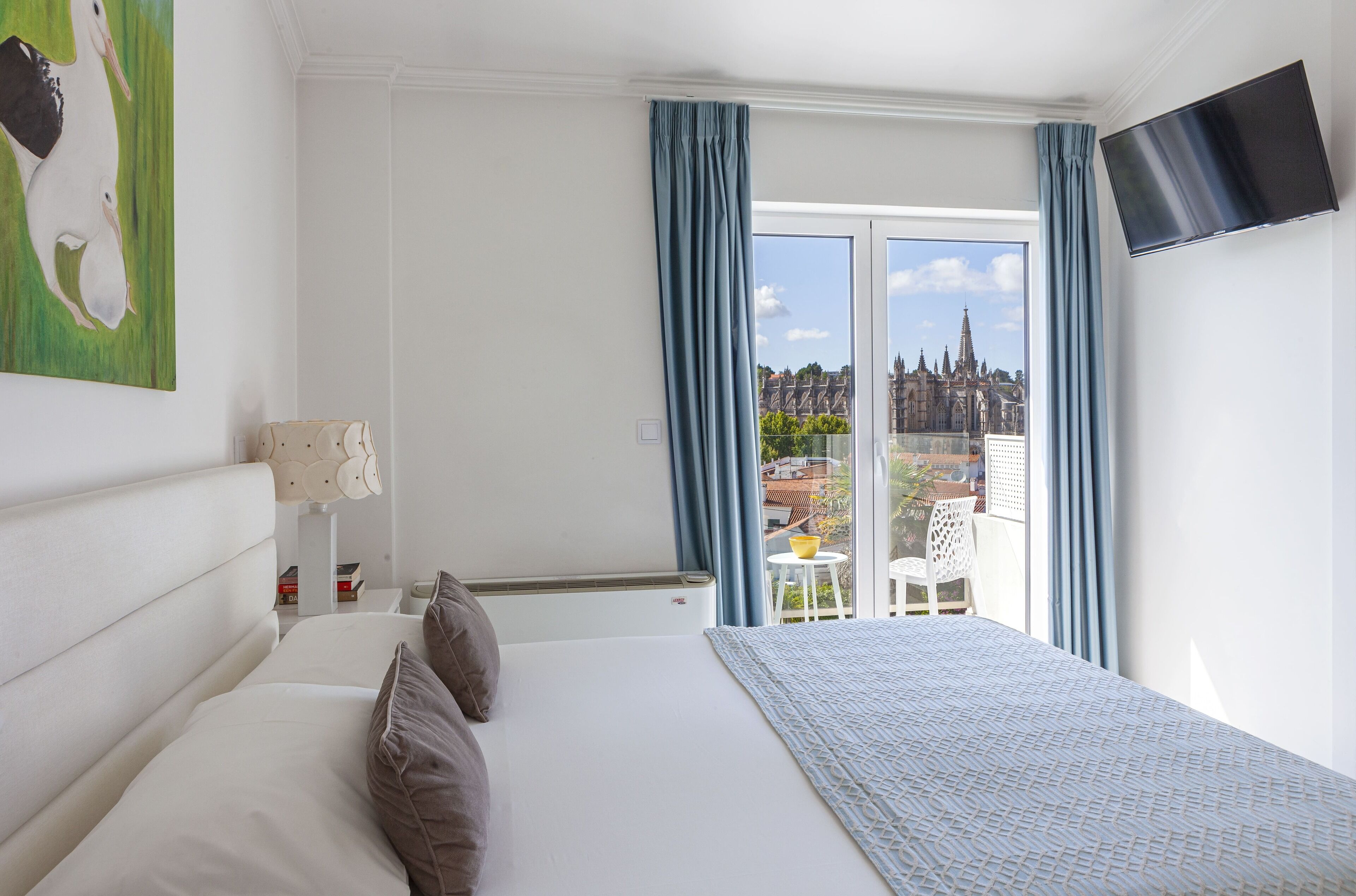 Habitación de hotel con una cama grande, un televisor de pantalla plana, un cuadro y vistas de la ciudad a través de una ventana con cortinas azules.