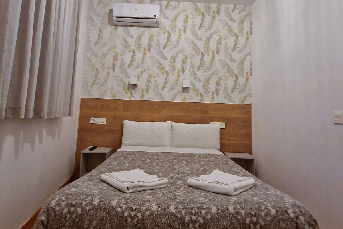 A hotel room with a bed, an air conditioning unit, a nightstand, and a remote control.
