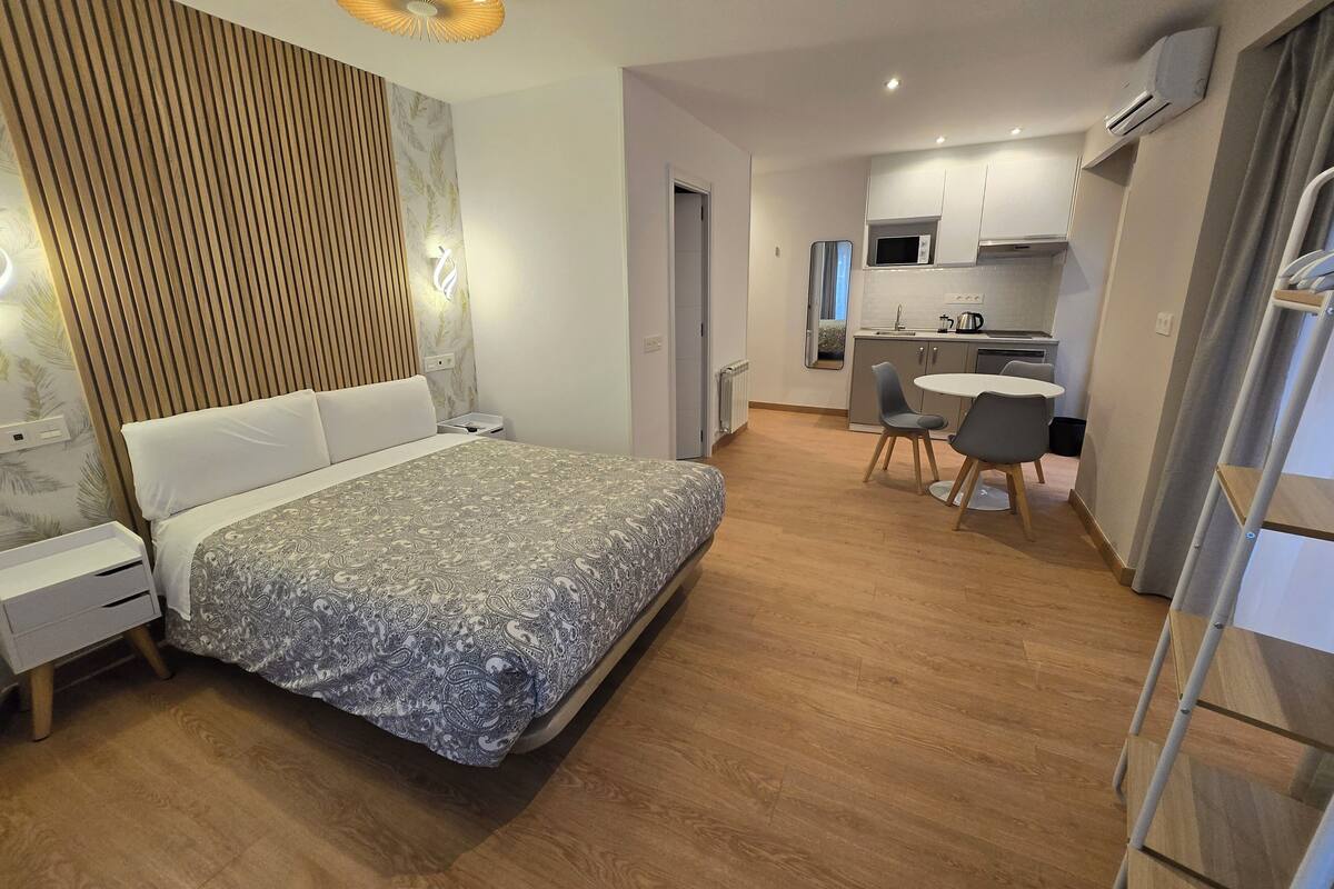 A modern hotel room with a large bed, bedside tables, a small desk, and a kitchenette.