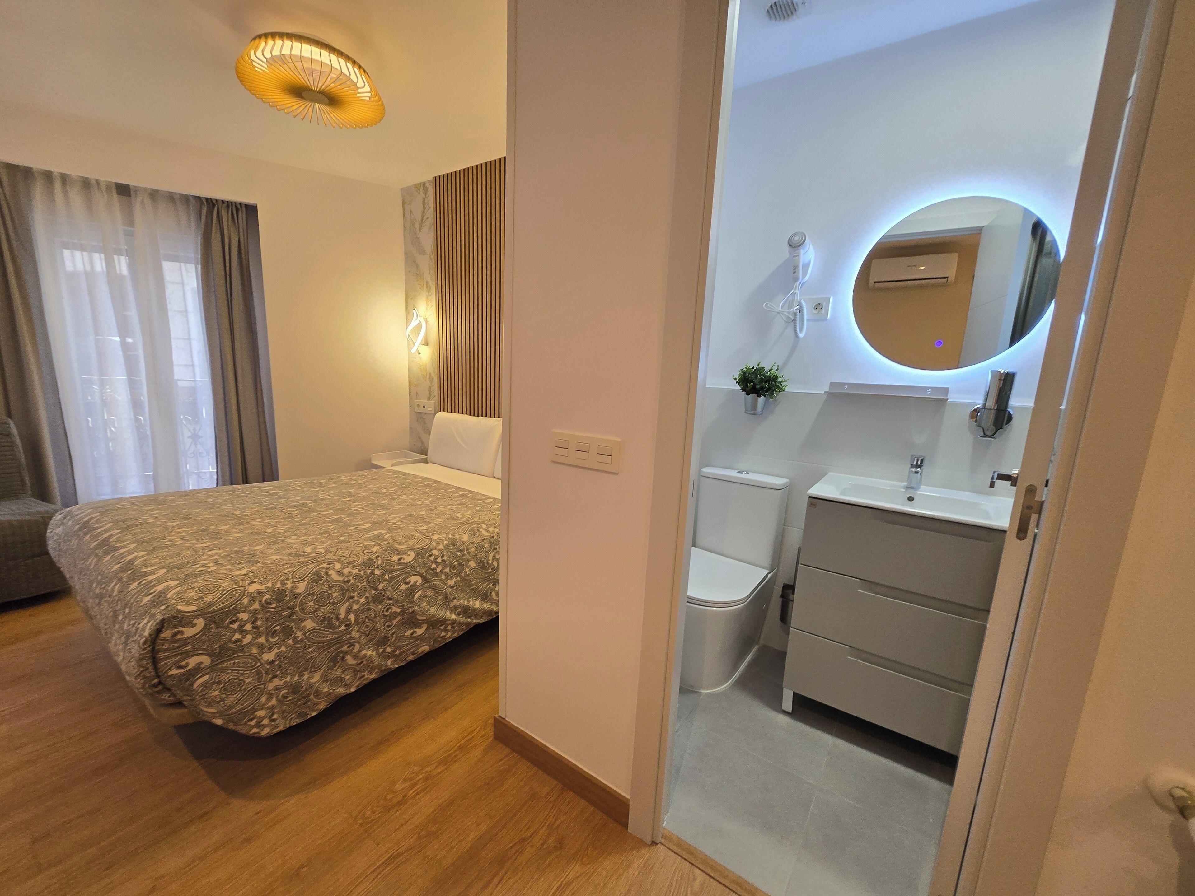 A modern bedroom with a patterned bedspread, a bedside lamp, and a view into a bathroom with a toilet, sink, and mirror.