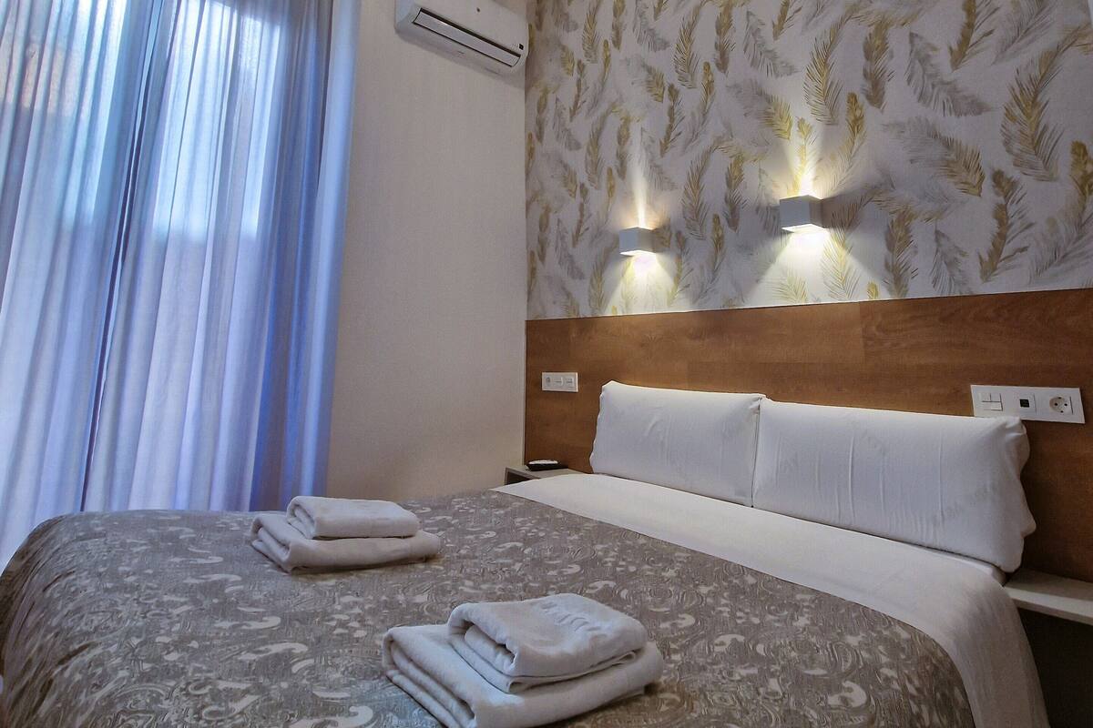 A hotel room with a large bed, a headboard with wall-mounted lights, an air conditioning unit, and a window with curtains.