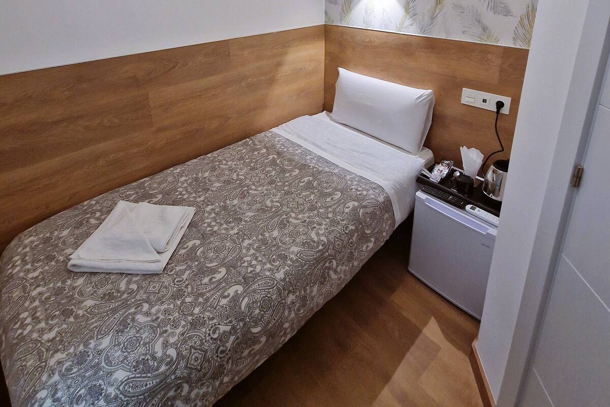 A small, neatly made bed with a patterned bedspread and a folded white towel. A small bedside table with a lamp and a few items is visible. The room has wooden paneling on the walls and a patterned wallpaper on one wall.