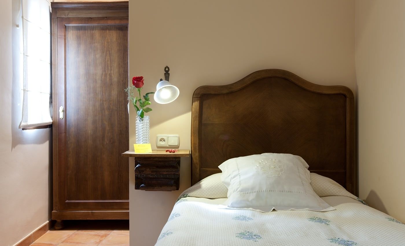 Un dormitorio con cabecera de madera, una mesita de noche con lámpara, un florero con una rosa roja y una cama con almohada blanca.