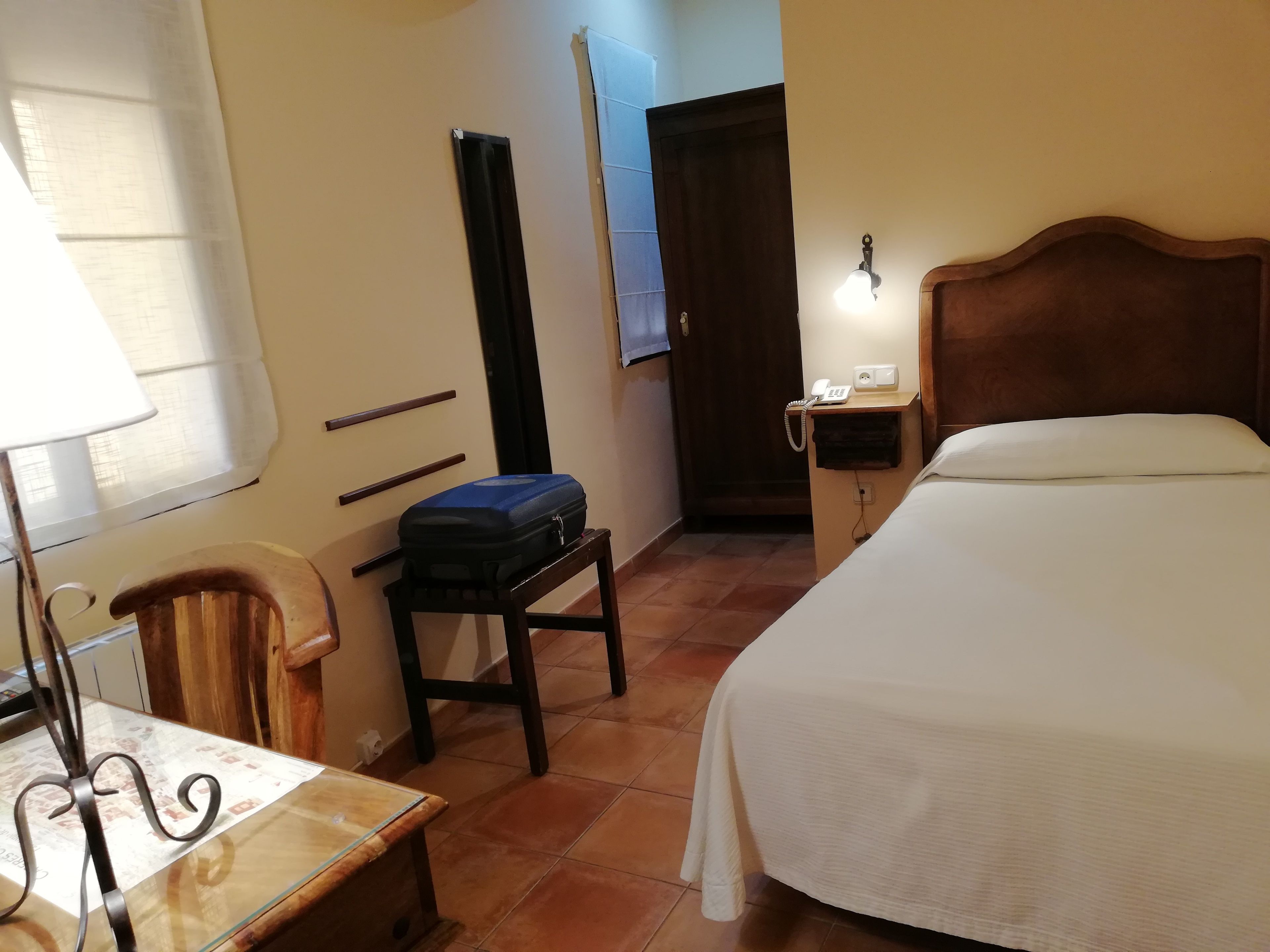 Una habitación de hotel con cama, escritorio con lámpara, una silla, maleta sobre una mesita, teléfono en la pared y una puerta.