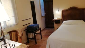 Una habitación de hotel con cama, escritorio con lámpara, una silla, maleta sobre una mesita, teléfono en la pared y una puerta.