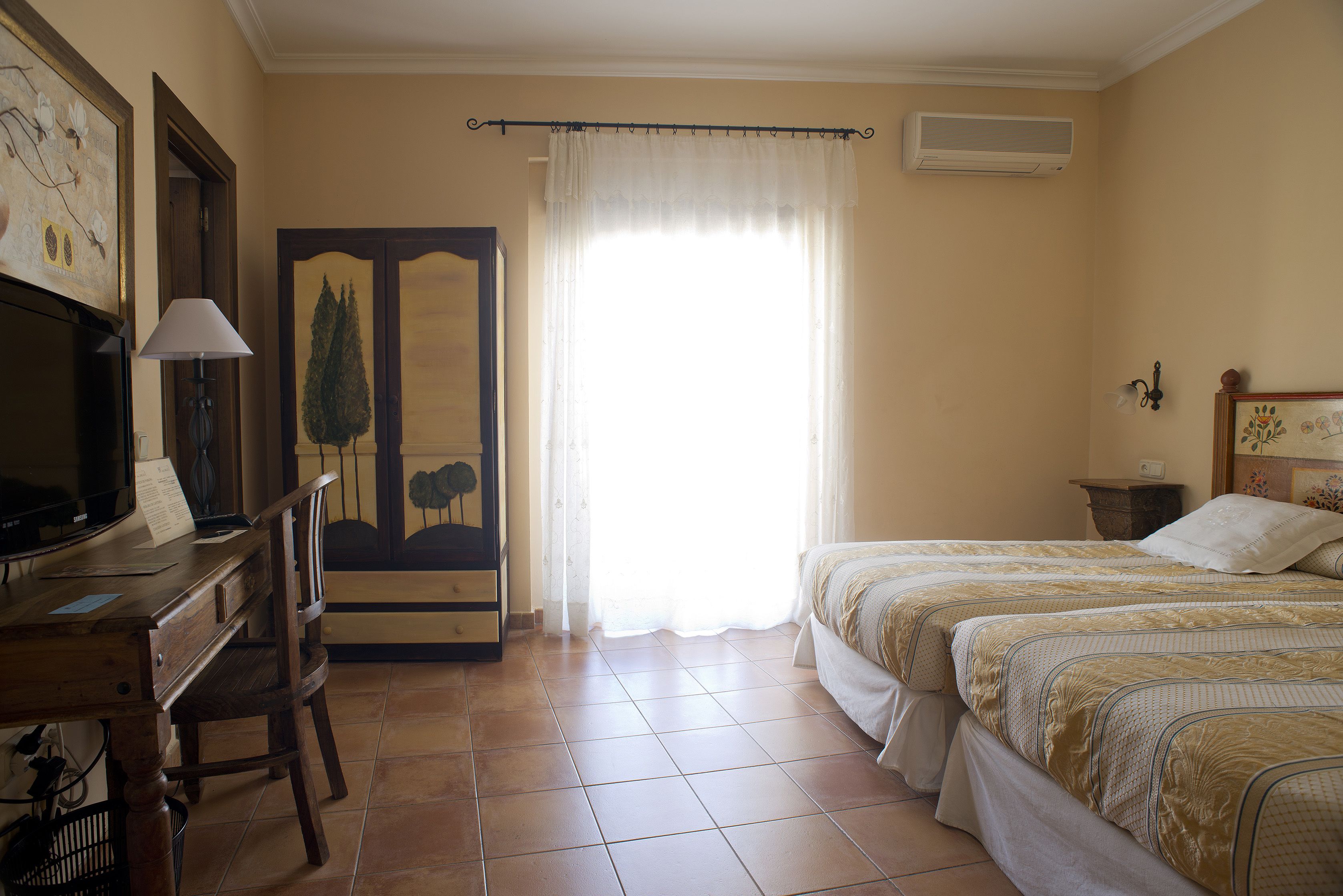 A bedroom with two beds, a desk with a lamp, a chair, a painting on the wall, and a window with curtains.