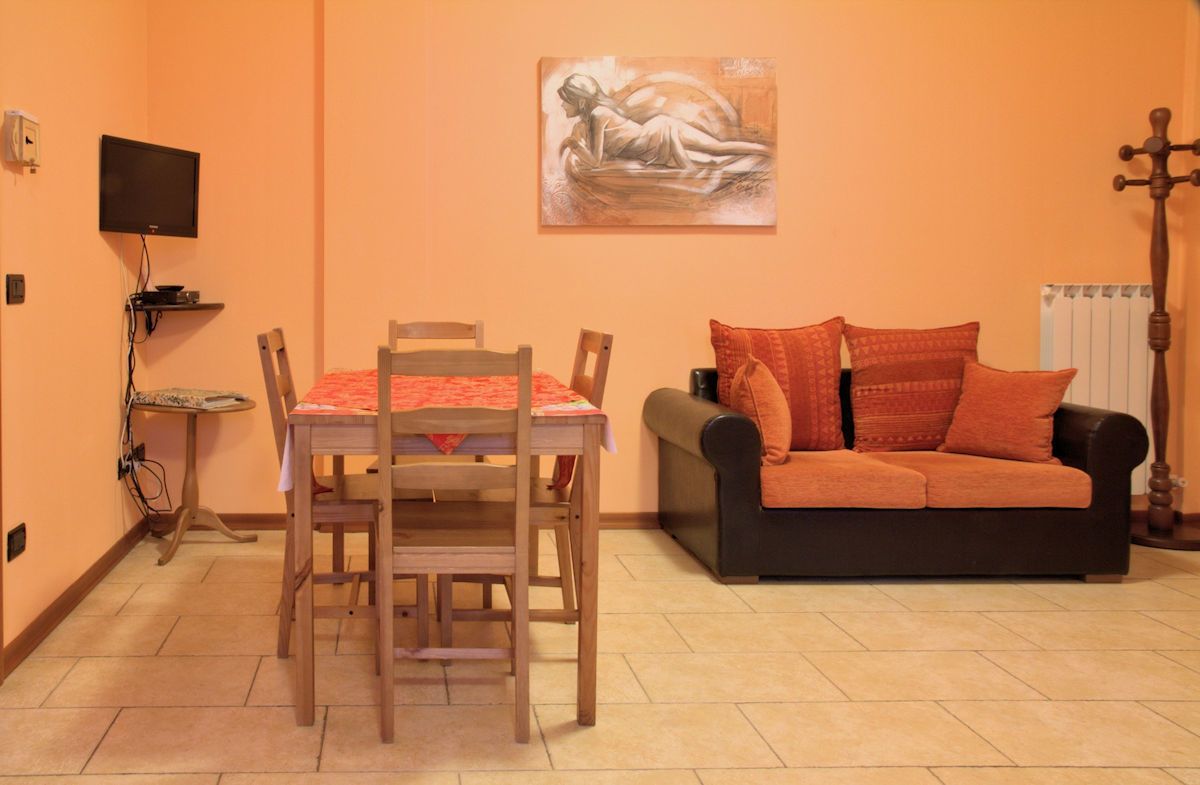 A room with a dining table, chairs, a black sofa with orange cushions, a painting on the wall, a television on a stand, and a radiator.