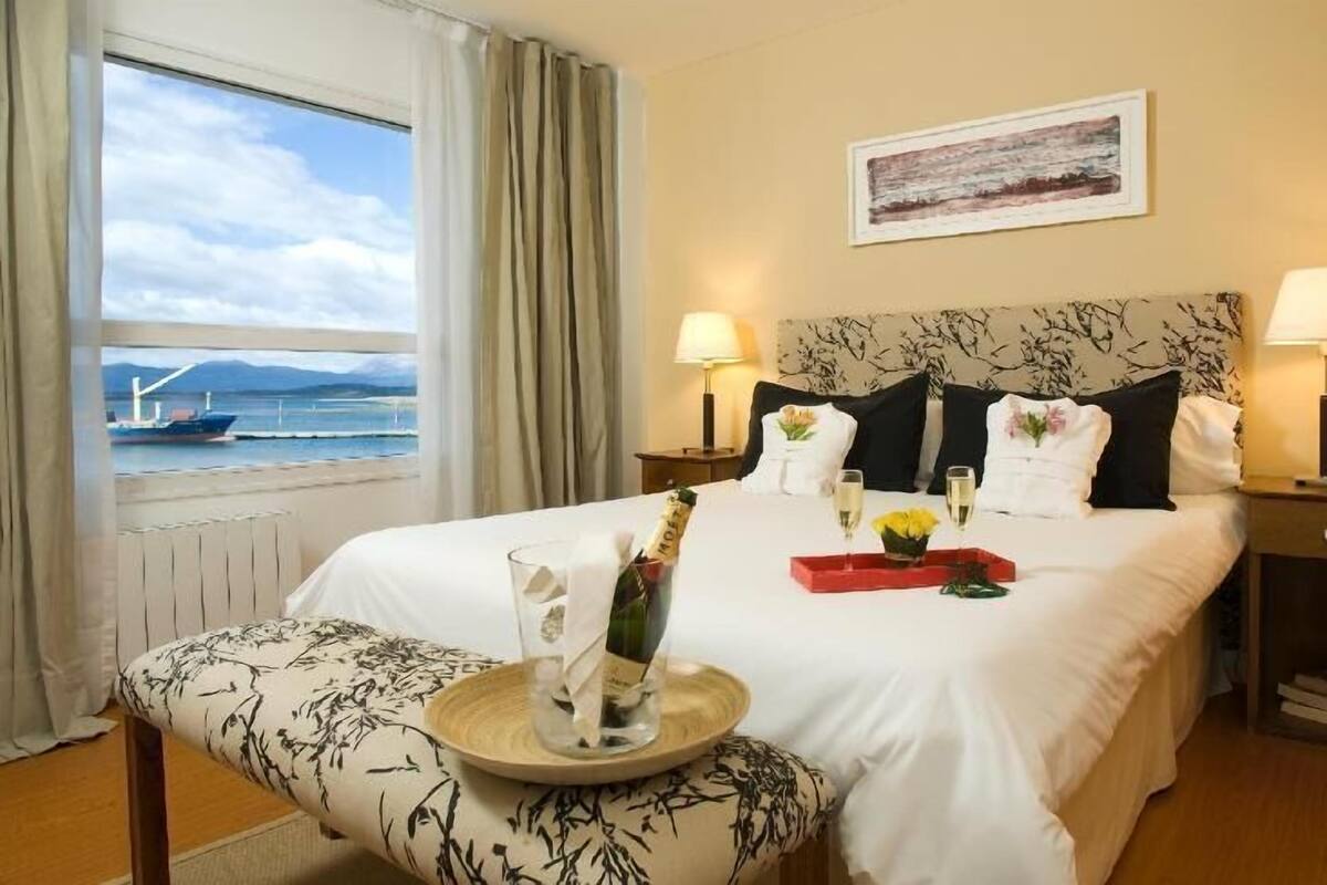 A hotel room with a bed, two bedside lamps, a window with a view of the sea, a framed artwork on the wall, and a small table with a bottle and glasses.