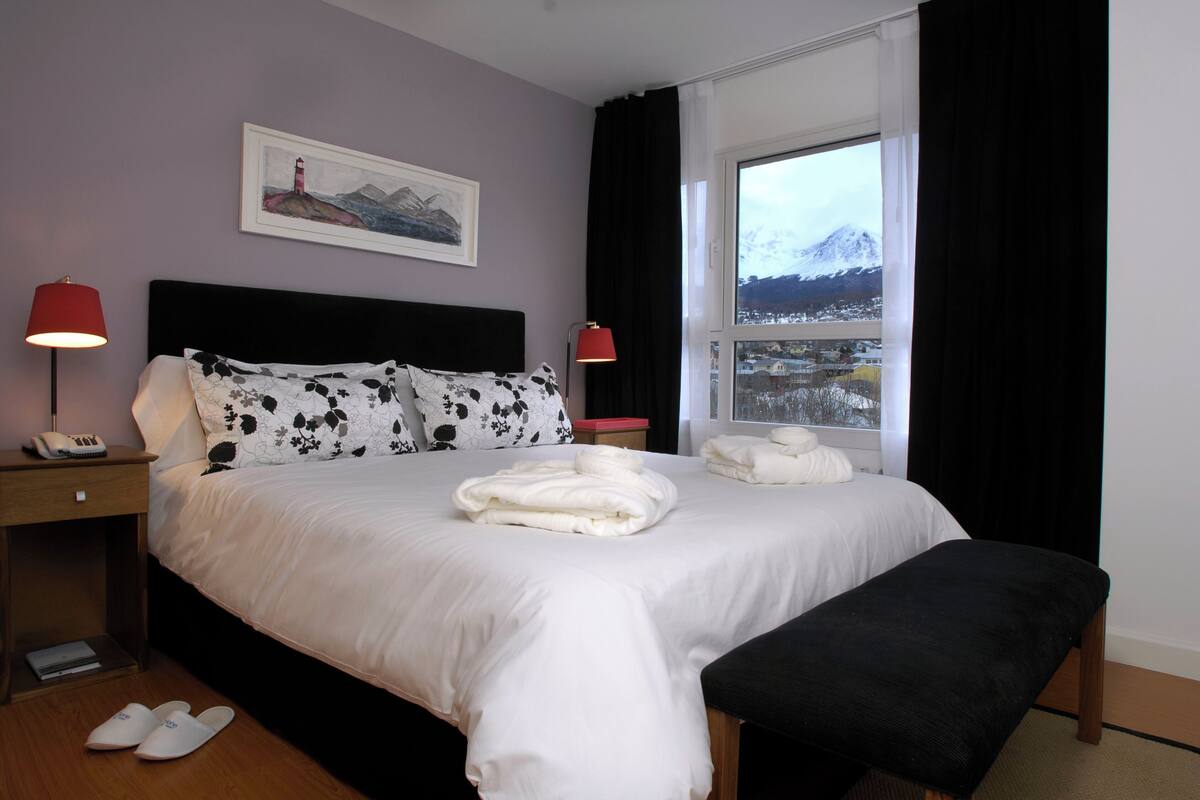 Un lit bien fait, avec du linge de lit blanc, une lampe de chevet, un tableau encadré au mur et, par la fenêtre, une vue sur une montagne enneigée.