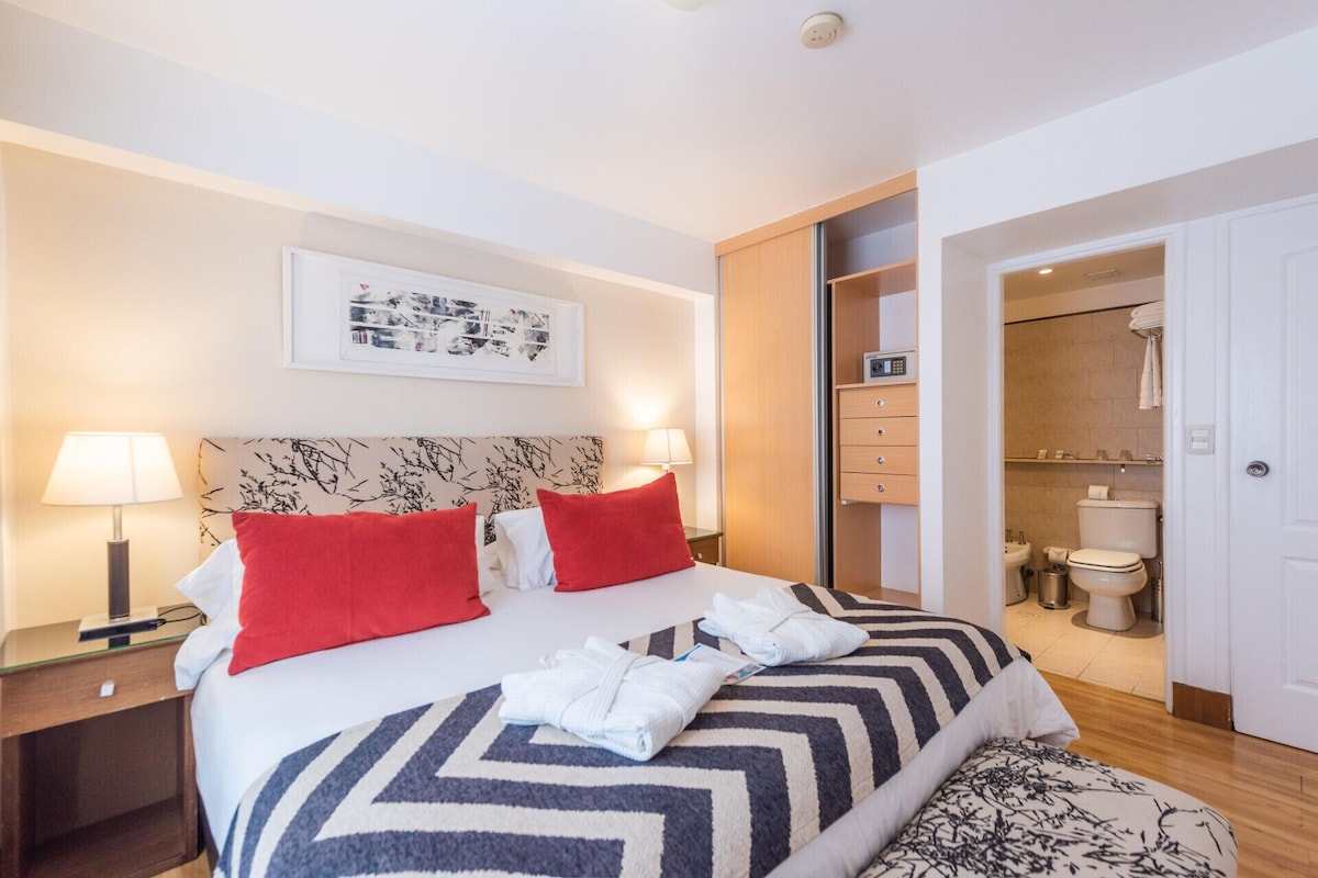 A hotel room with a bed, two red pillows, a bedside table with a lamp, a wall-mounted picture, a bathroom with a toilet and sink, and a walk-in closet.