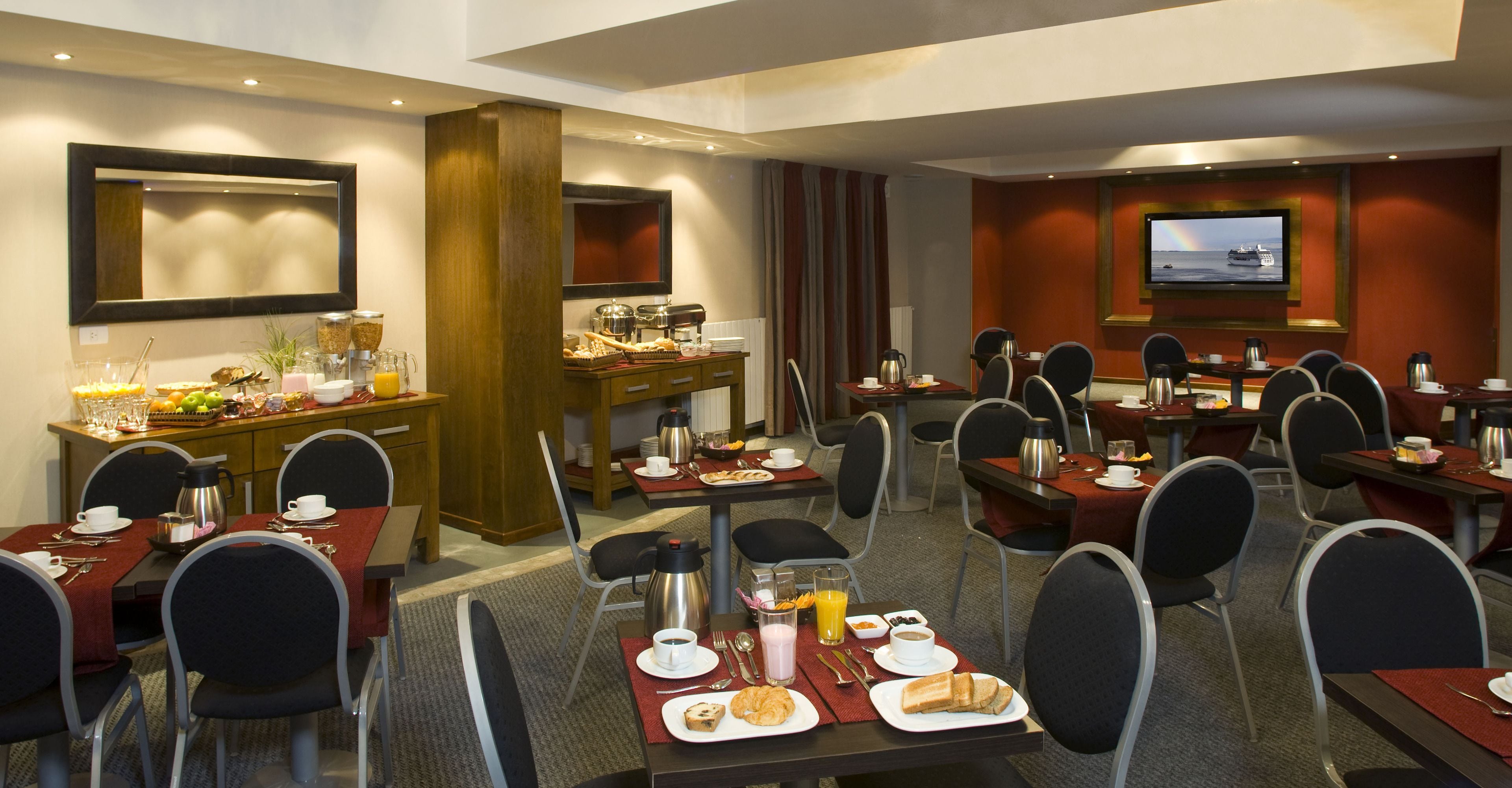 Une salle à manger d’hôtel avec plusieurs tables, des chaises, un buffet pour le petit-déjeuner et un téléviseur qui diffuse des images d’un navire.
