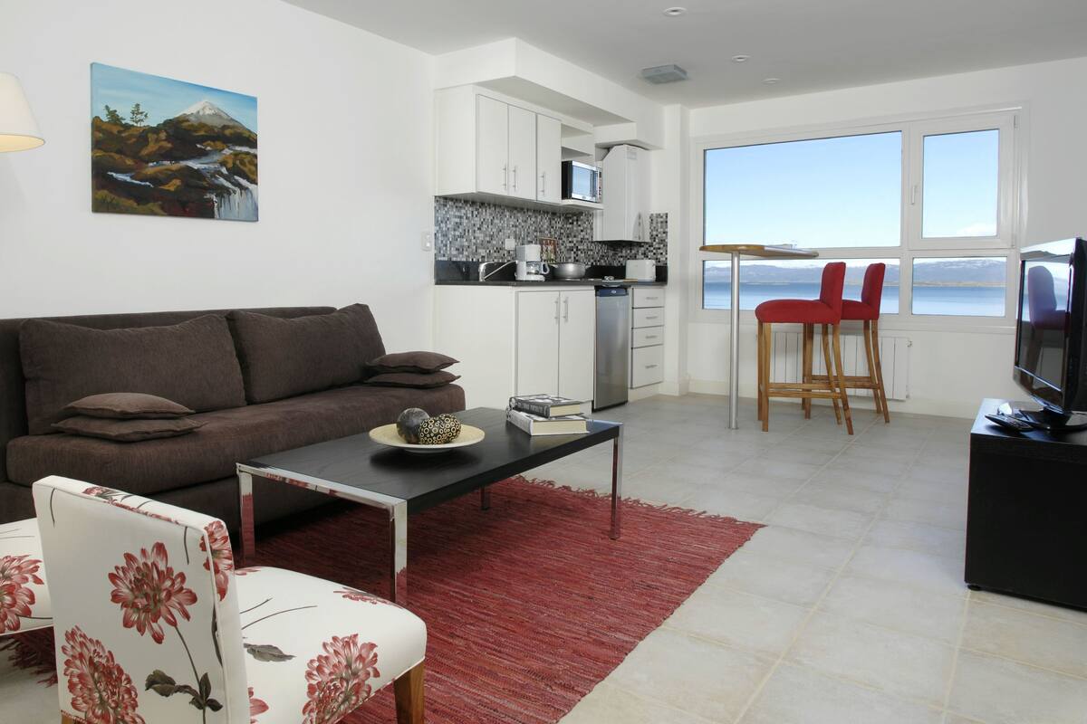 A modern living room with a sofa, coffee table, and a TV. There's a kitchen area with cabinets and a view of the sea through large windows.