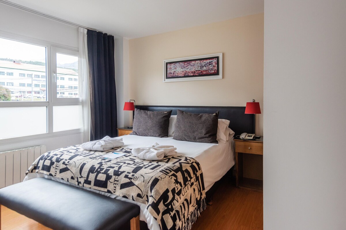 A hotel room with a bed, bedside lamps, a window with curtains, a wall-mounted painting, and a bench.