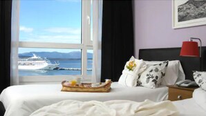 A hotel room with a bed, a nightstand, a red lamp, and a window with a view of a cruise ship.