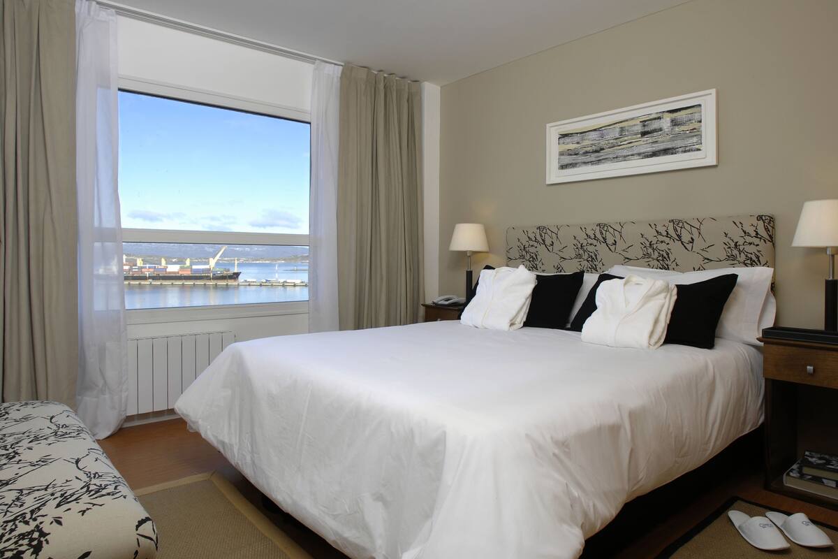 A hotel room with a large bed, two bedside lamps, a nightstand, a framed picture on the wall, and a view of a harbor with ships.