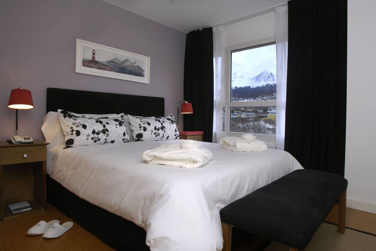 A neatly made bed with white linens, a bedside lamp, a framed picture on the wall, and a window with a view of a snowy mountain.
