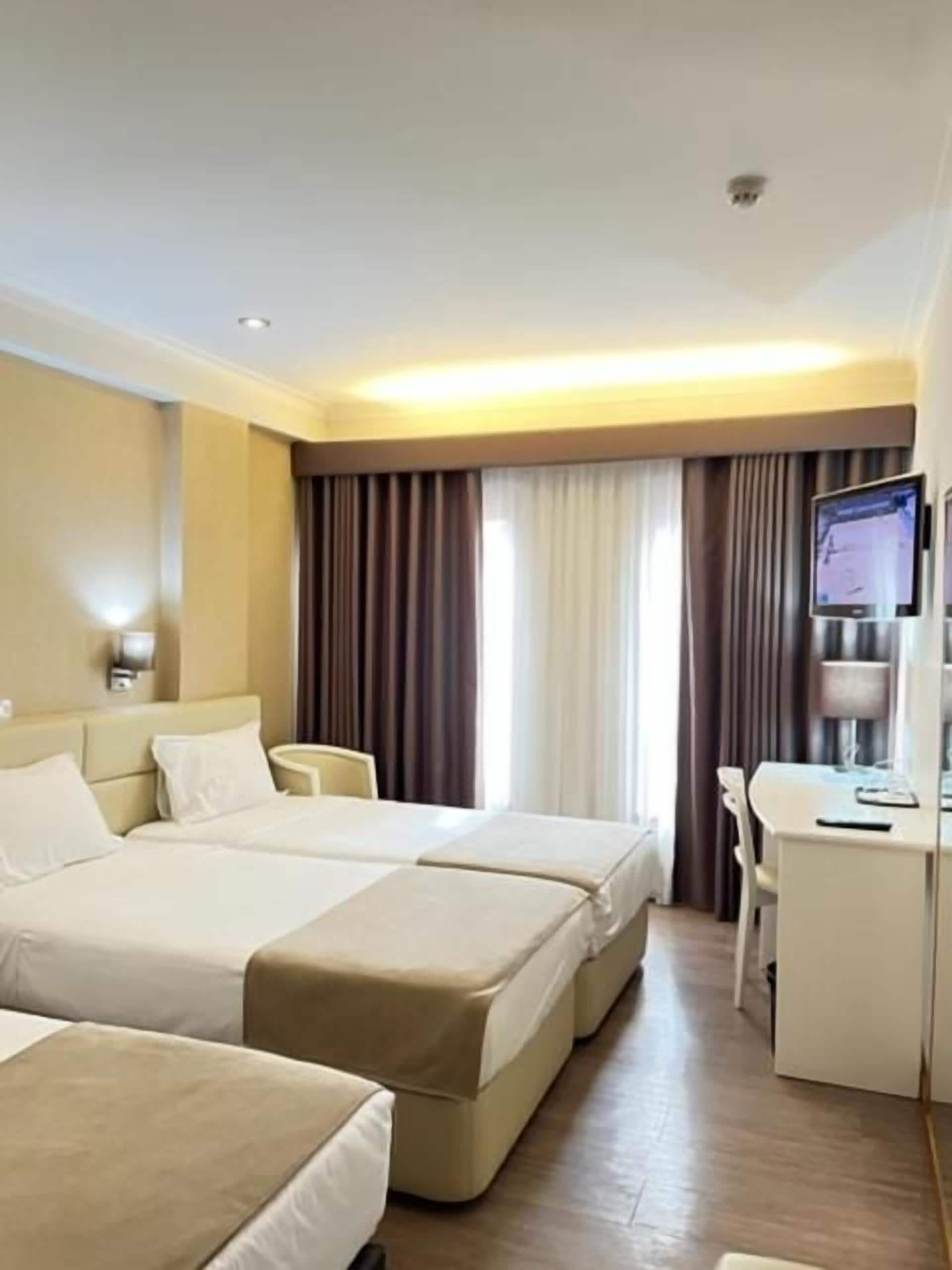A hotel room with two beds, a desk, a TV, and a window with curtains.