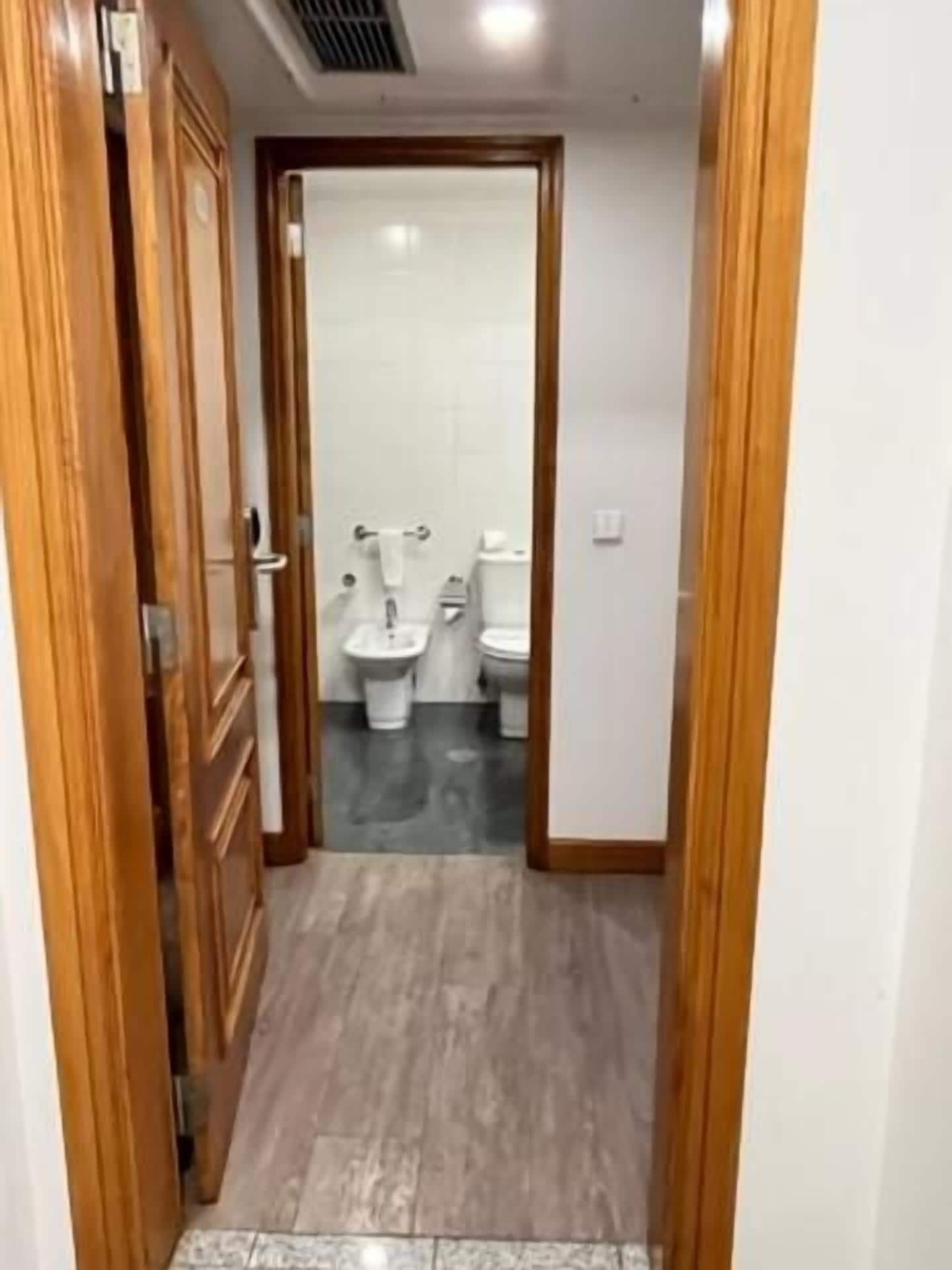 A narrow hallway leading to a bathroom with two toilets and a sink.