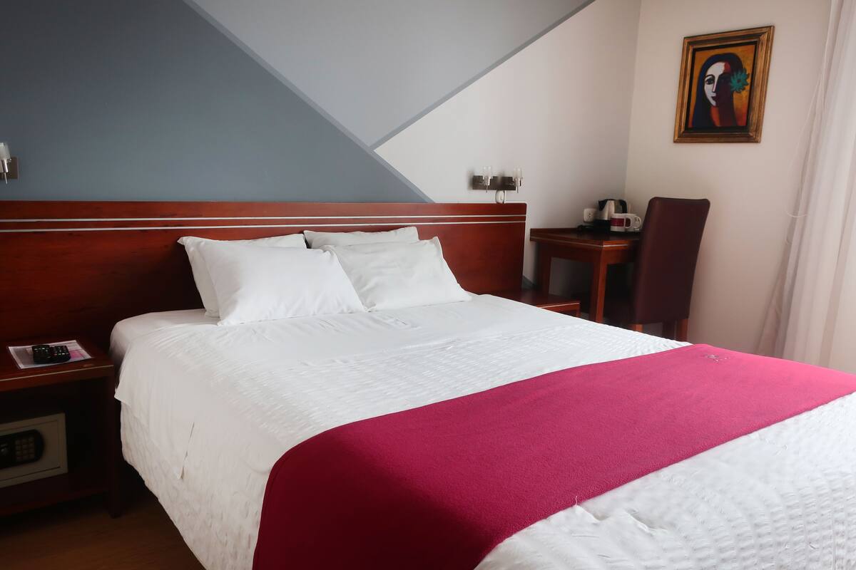 A hotel room with a bed, a nightstand, a table with a chair, and a painting on the wall.