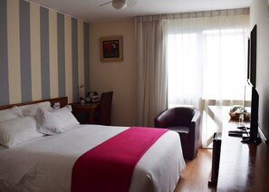 Quarto de hotel com cama, escrivaninha, cadeira, televisão e janela com cortinas.