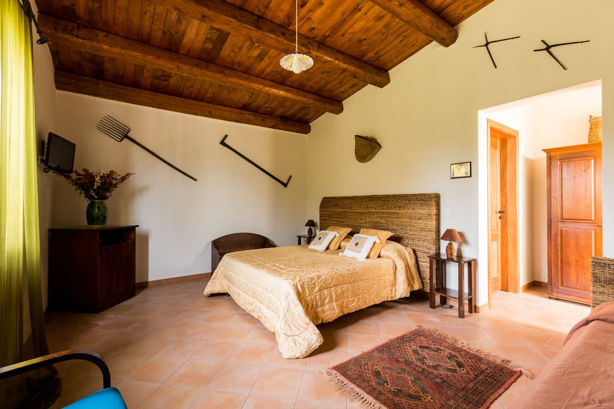 Une chambre avec un plafond en bois, un lit recouvert d’une couverture beige, une petite table avec une lampe et une chaise.