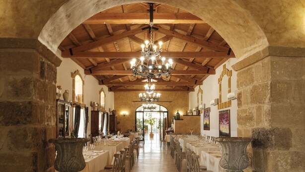 Una sala alargada y estrecha, con techos altos y paredes de piedra, que cuenta con varias mesas dispuestas para comer.