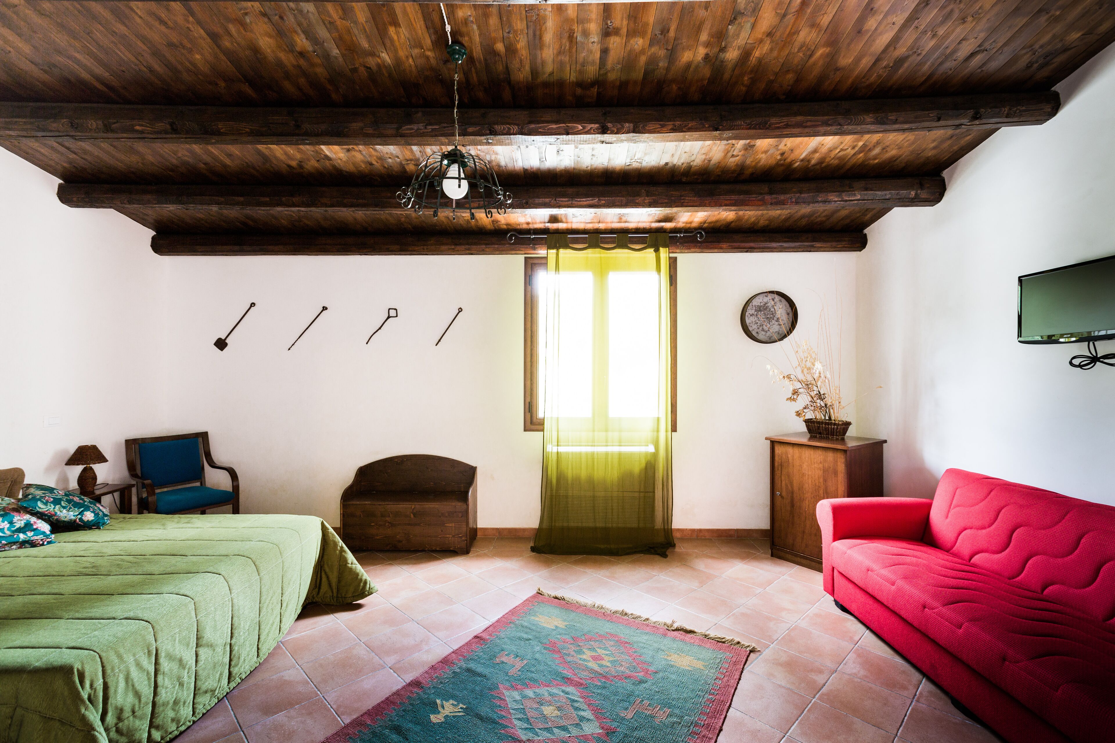 Una stanza con soffitto in legno, un letto con copriletto verde, un divano rosso, un piccolo armadio in legno e un televisore appeso al muro.
