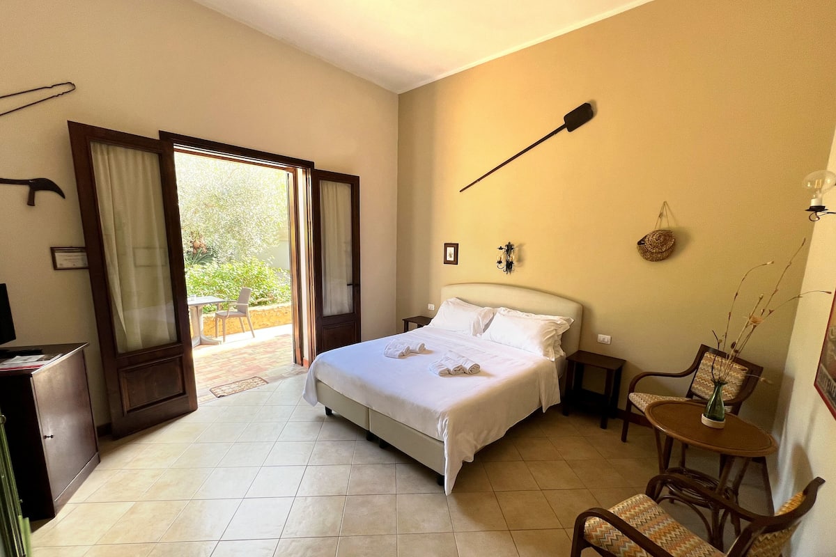 Une chambre d’hôtel comprenant un lit, une chaise, une petite table, une lampe fixée au mur et une porte donnant sur un espace extérieur.