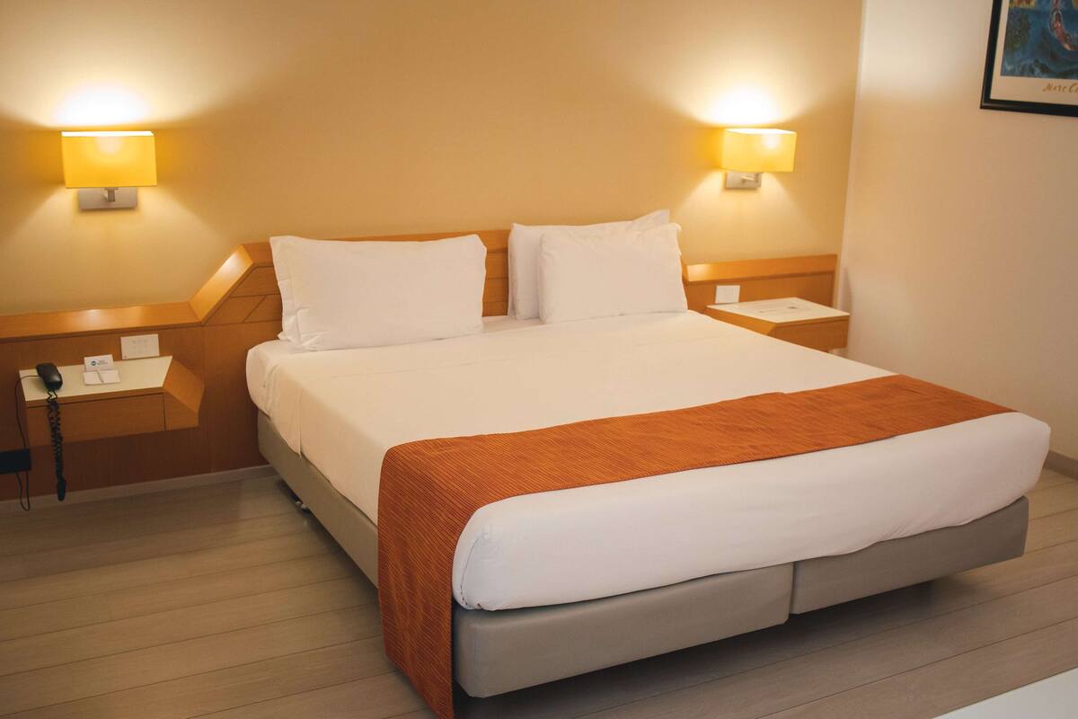 Een keurig opgemaakt bed met witte lakens en een bruine plaid, met aan beide zijden nachtkastjes met lampen, in een hotelkamer met houten vloer.