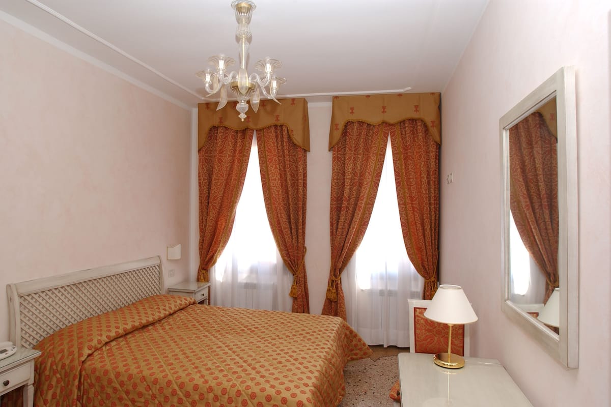 Une chambre d’hôtel avec un lit double, une table de chevet avec une lampe, un miroir et deux fenêtres avec des rideaux.