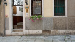 Une façade d’immeuble avec une porte, une fenêtre ornée de barreaux en métal et une jardinière remplie de fleurs roses.