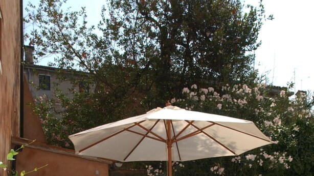 Une terrasse extérieure avec une table et des chaises sous un grand parasol, entourée de murs en briques et de plantes en fleurs.